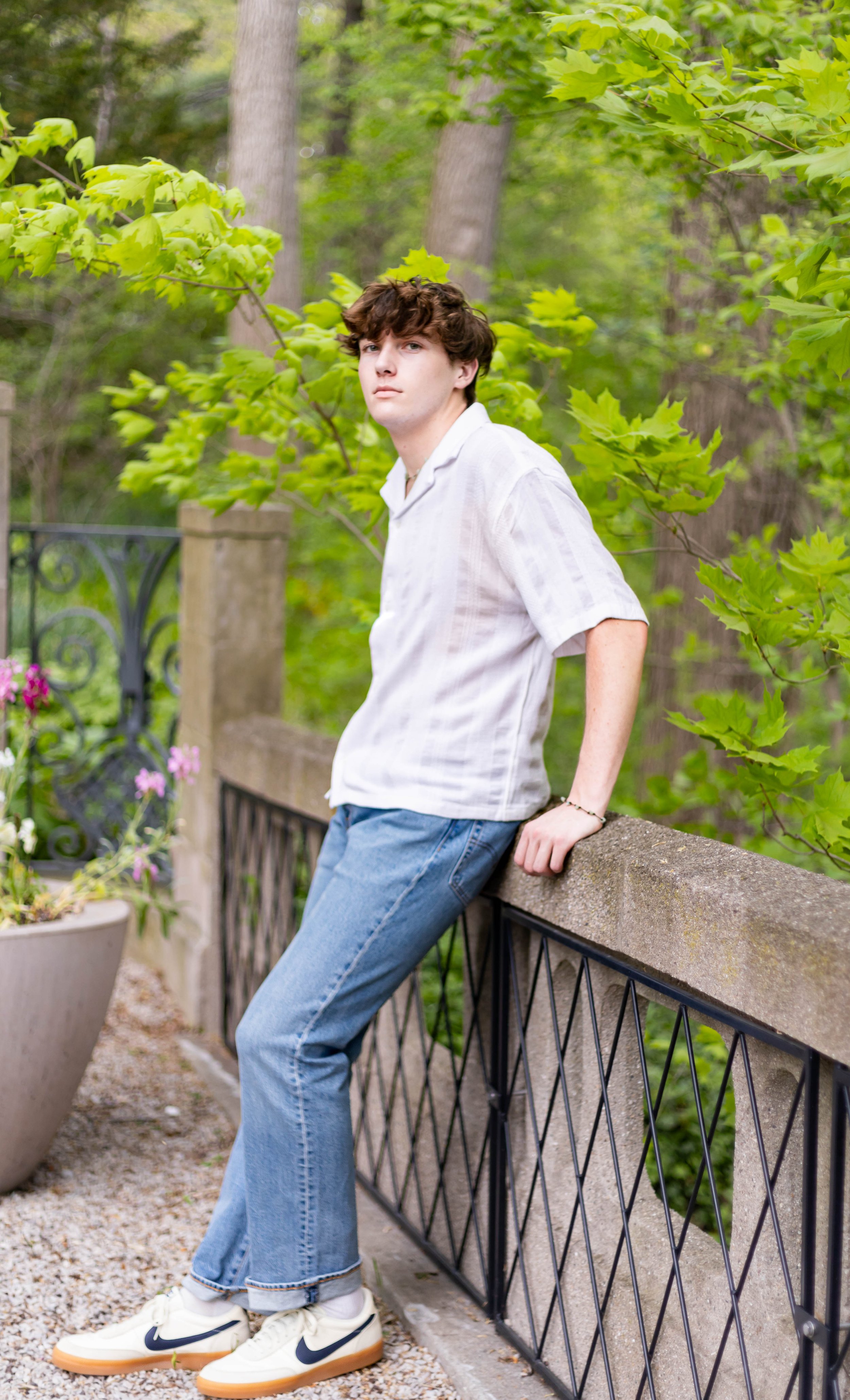A young man with brown hair wearing a white shirt, blue jeans, and white sneakers with black swooshes, leaning against a stone and metal railing in a lush, green outdoor setting.