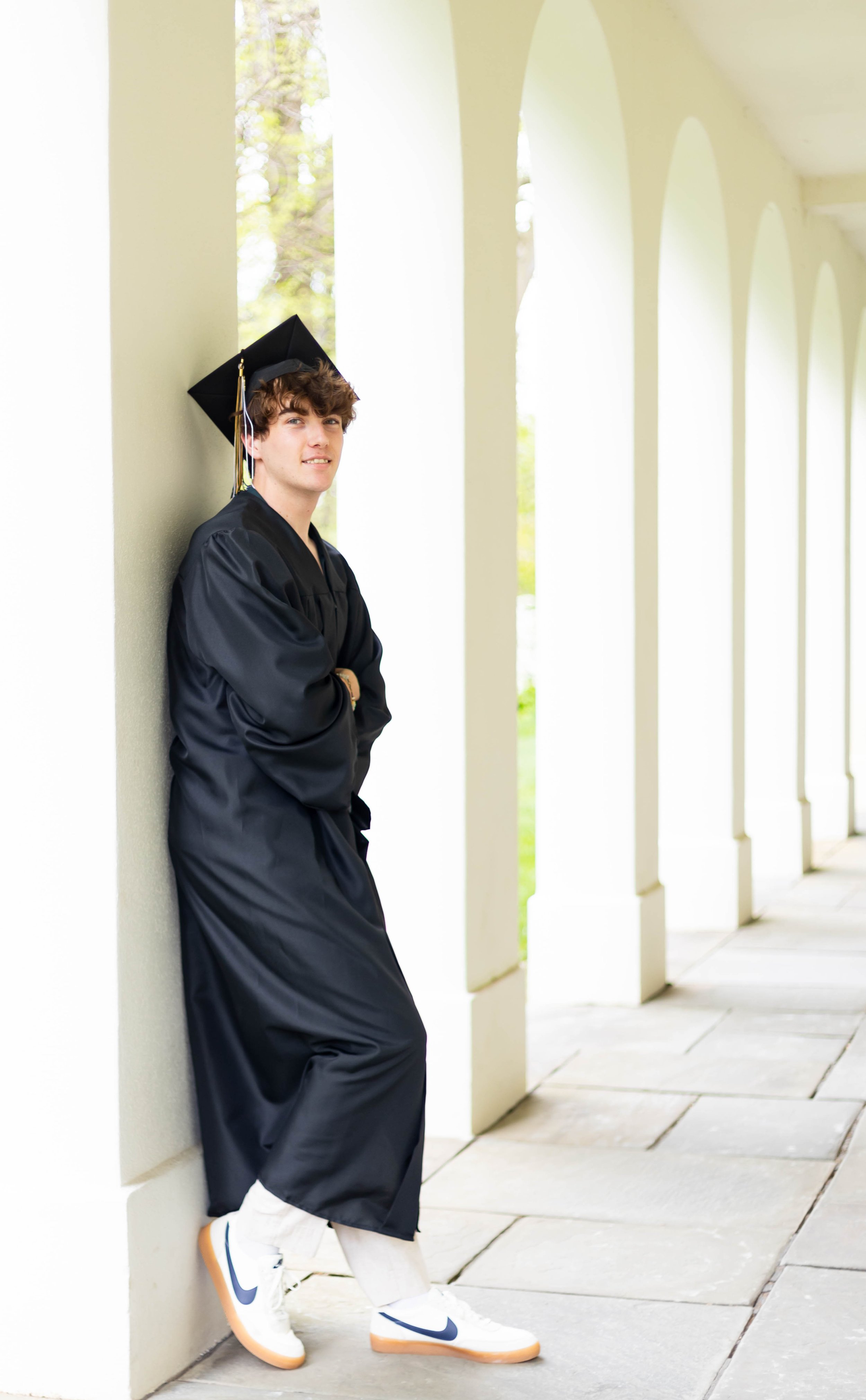 Young man in a black graduation gown and cap leaning against a white wall with archways, wearing white Nike sneakers with navy swoosh, outdoors during daytime.