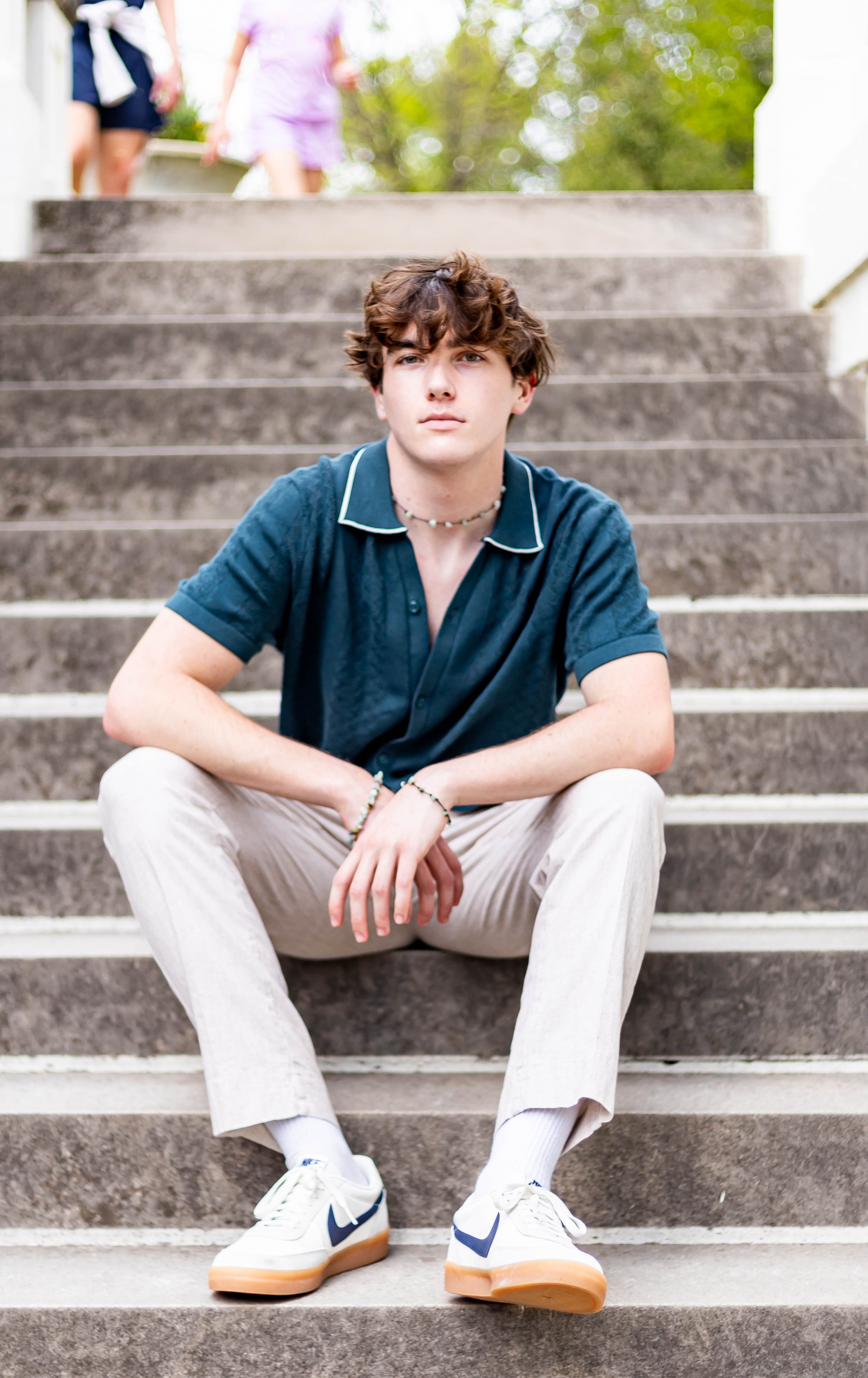 A young man with curly brown hair wearing a dark blue short-sleeve shirt, beige pants, and white sneakers, sitting on outdoor stairs with greenery in the background.