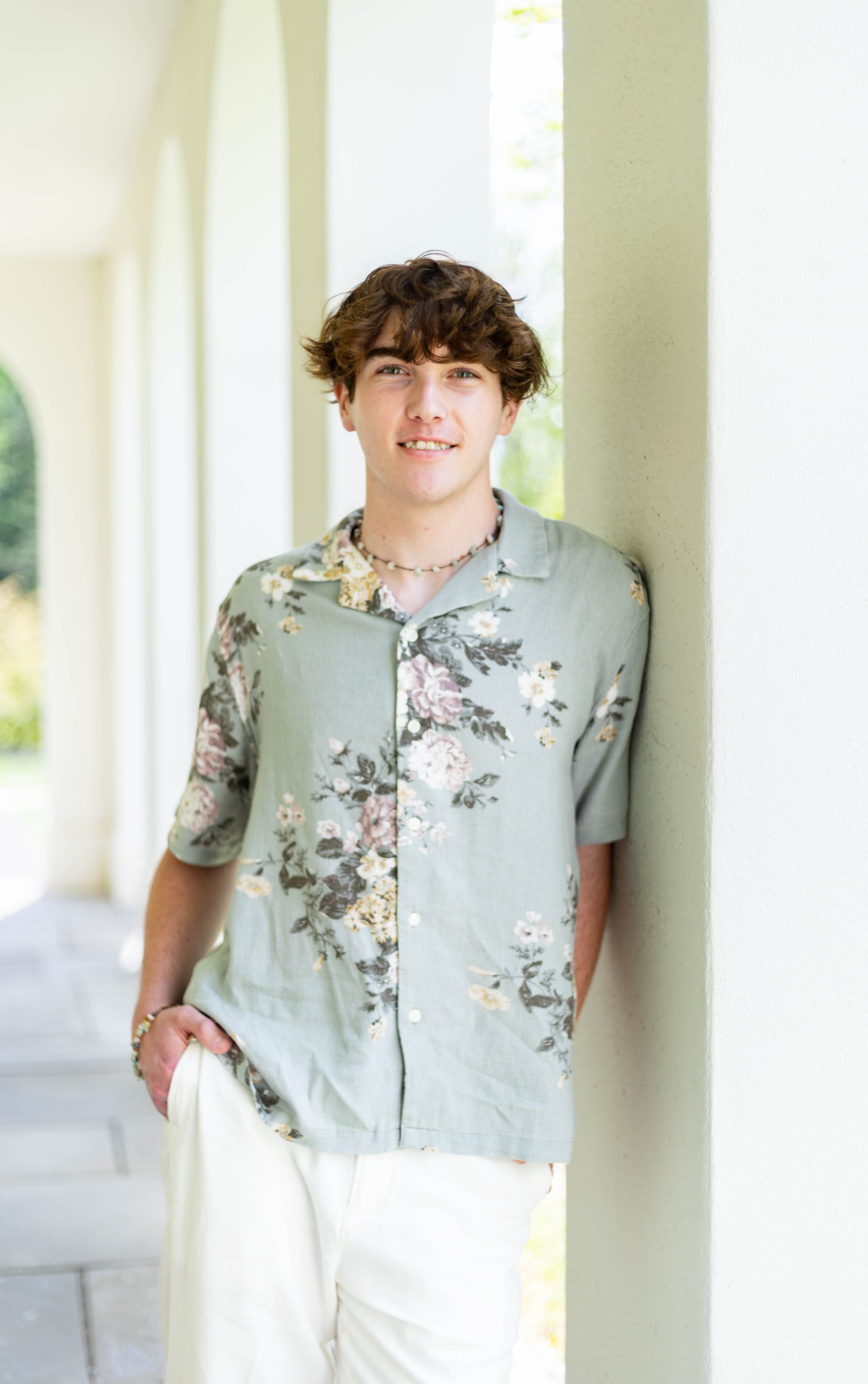Young man with curly brown hair smiling, dressed in a floral short-sleeve shirt and white pants, standing outdoors next to a beige wall