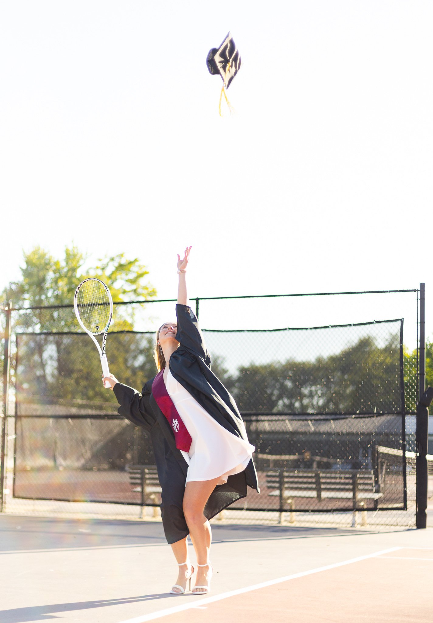 A woman in a graduation gown and heels on a tennis court throws her cap into the air.