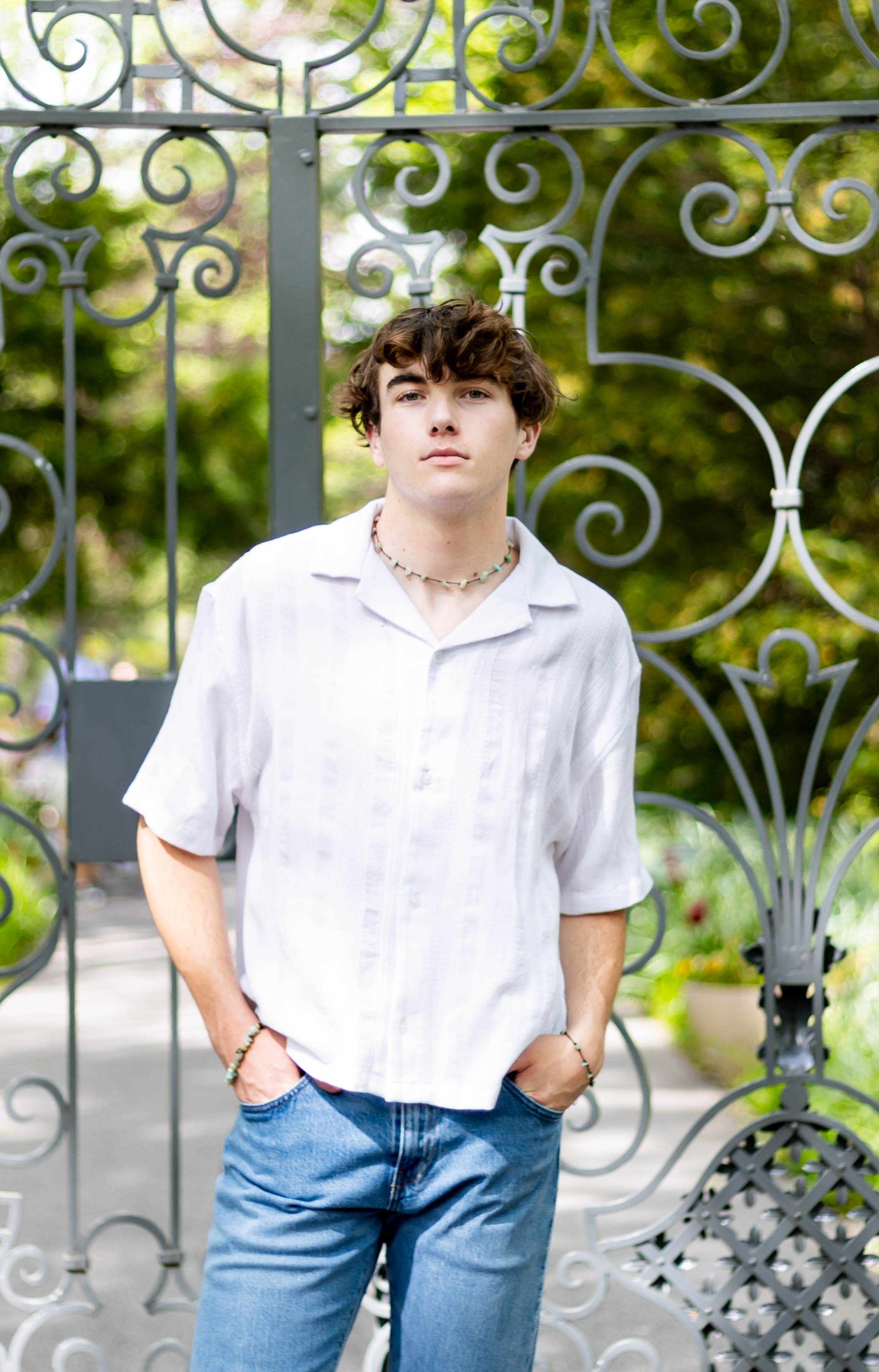 Young man with brown curly hair and fair skin standing outdoors in front of a decorative metal gate with green foliage in the background. He wears a white button-up shirt, blue jeans, and jewelry including necklaces and bracelets.