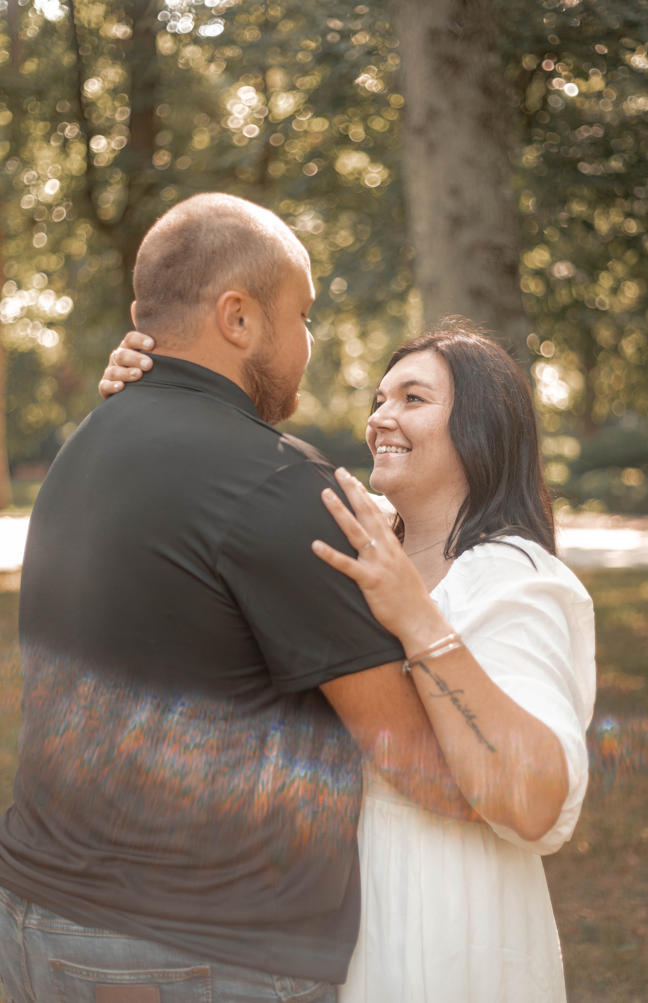 A couple embraces in a park, gazing into each other's eyes, with trees and warm sunlight in the background.