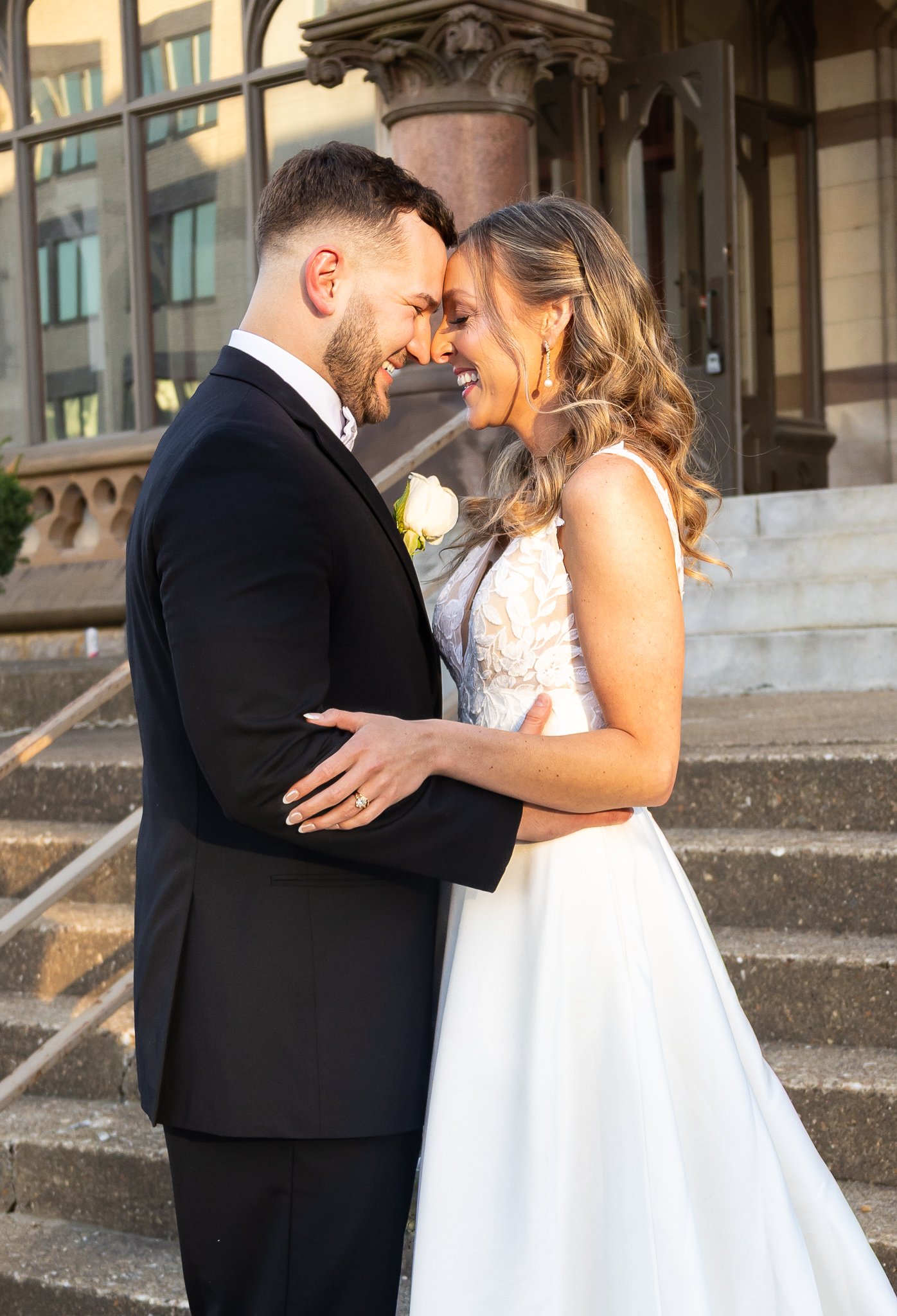 A bride and groom sharing a moment outside on steps, smiling with their foreheads touching. The groom is wearing a black tuxedo and the bride is in a white wedding dress with lace details.