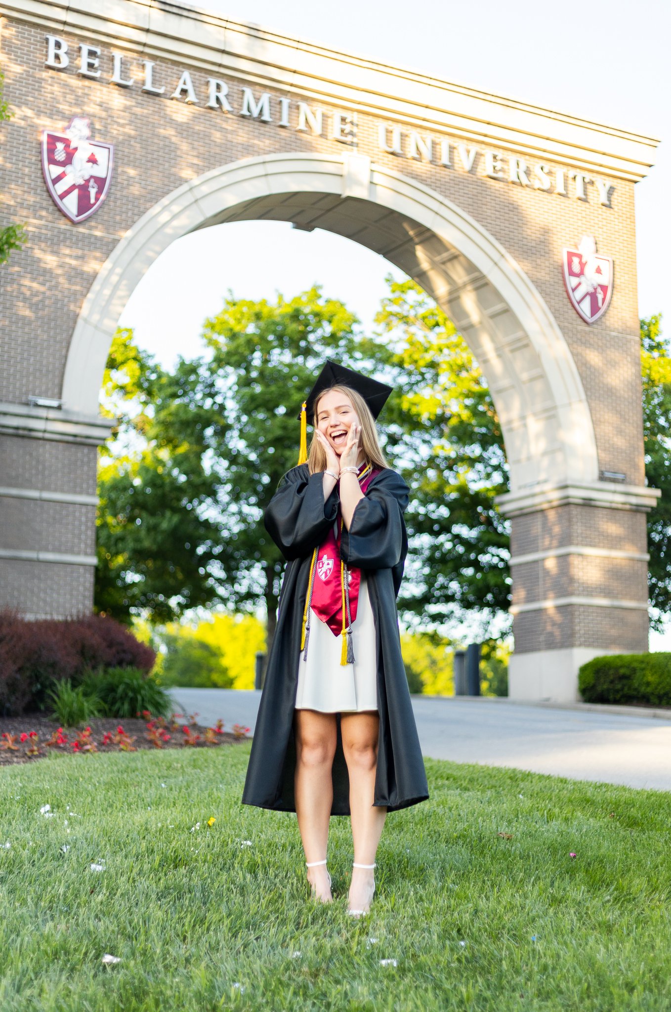 A young woman in a graduation gown and cap standing on grass in front of the Belarmine University arch, smiling and celebrating her graduation.