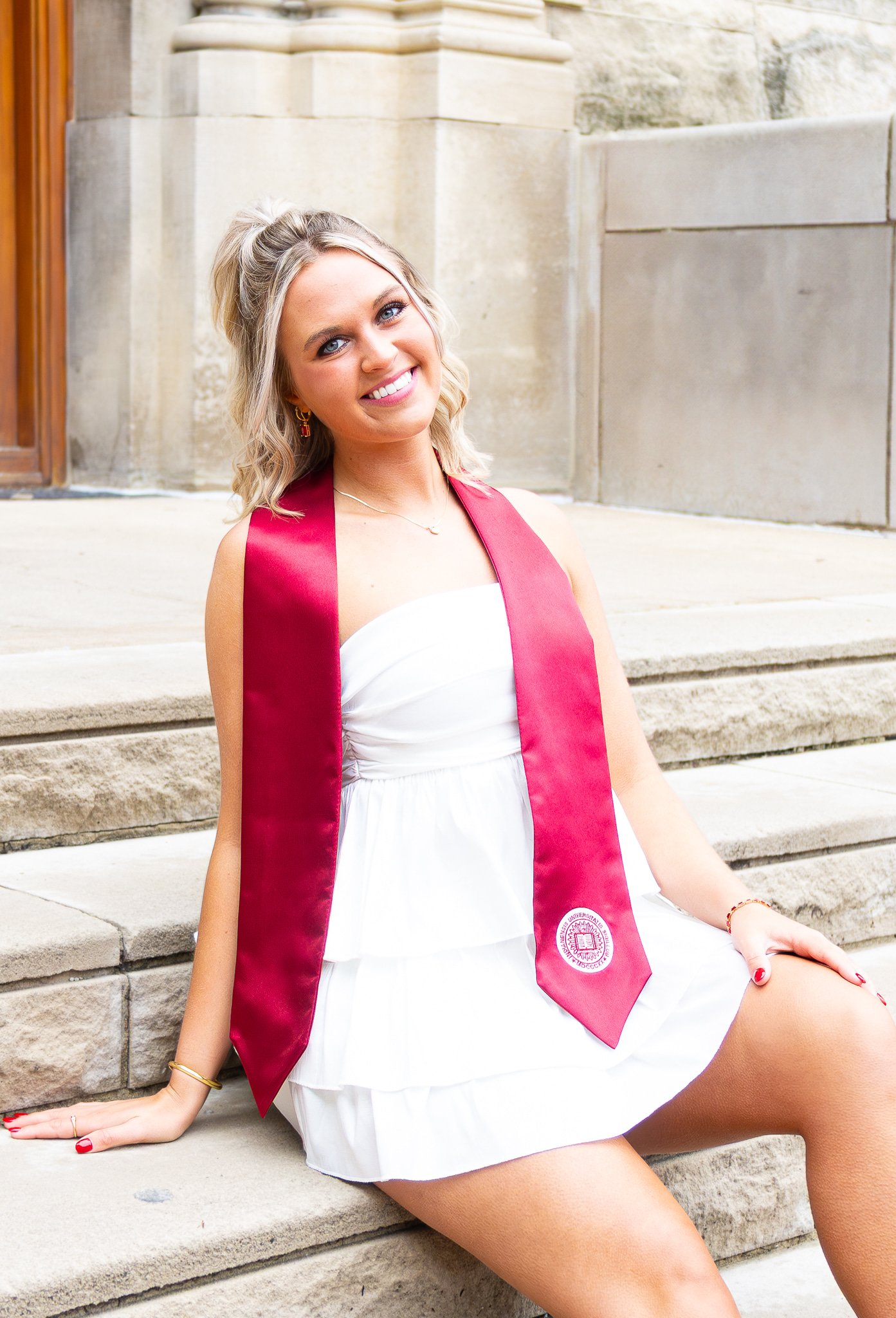 Smiling young woman sitting on steps in a white dress with a red graduation stole outside a stone building.