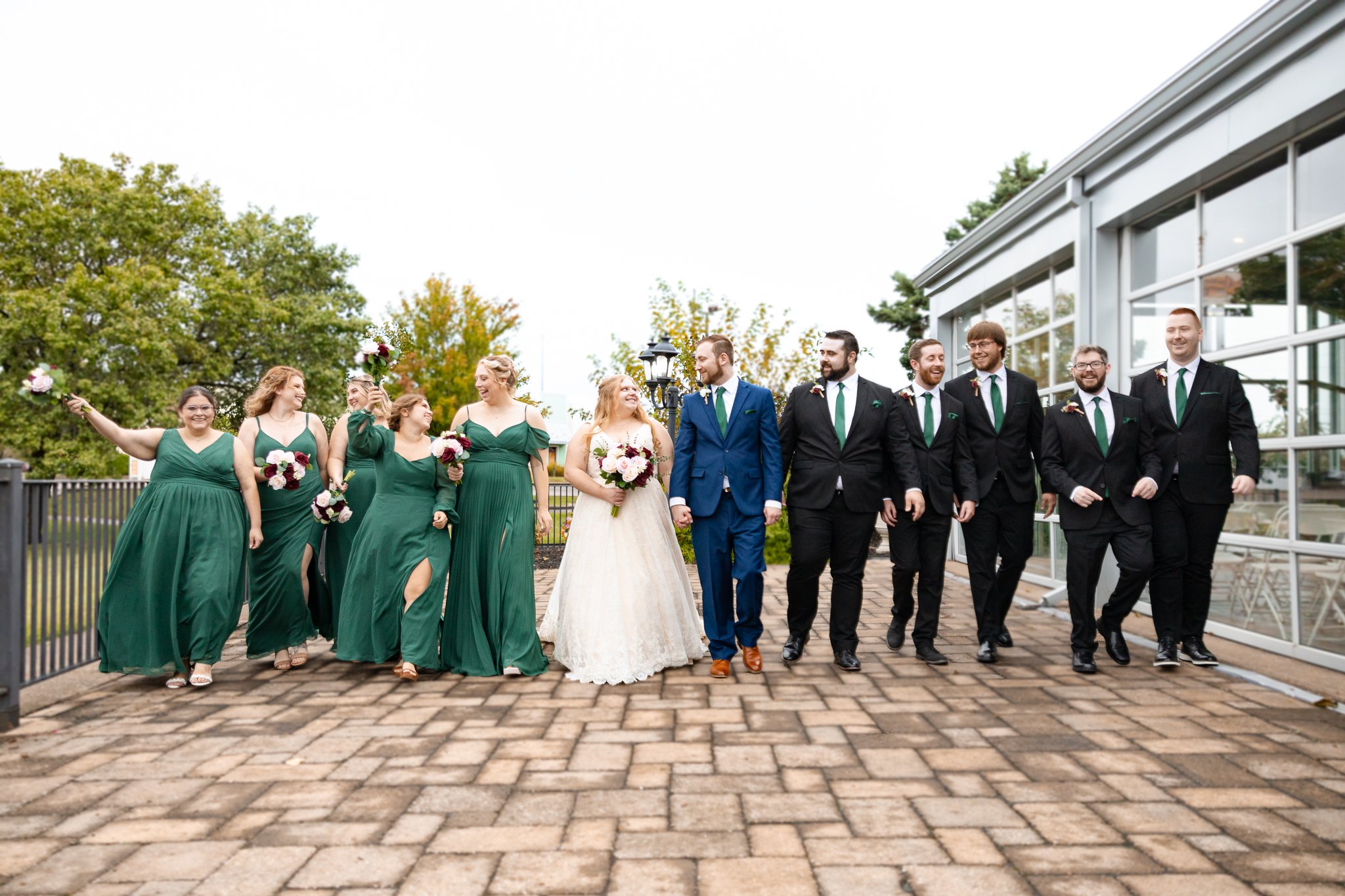 Bride and groom walking with wedding party on brick pathway, women in green dresses, men in black suits with green ties, bride in white wedding dress holding bouquet, groom in blue suit, group smiling and enjoying outdoor wedding celebration.