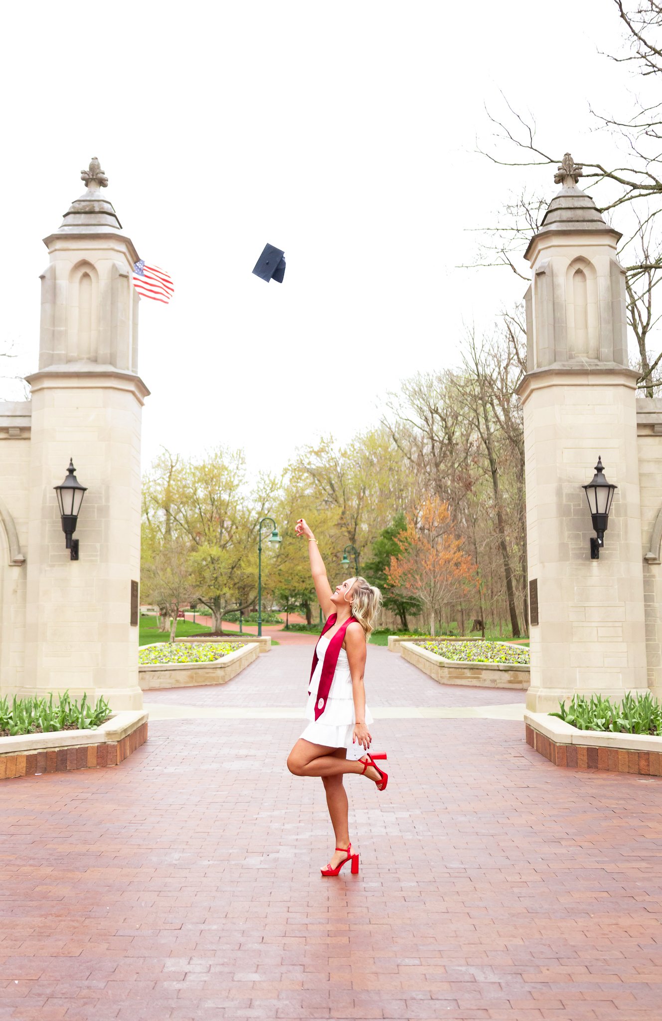 A woman in a white dress and red high heels is tossing her graduation cap into the air at an outdoor graduation ceremony. She is standing between two stone pillars with an American flag and trees in the background.