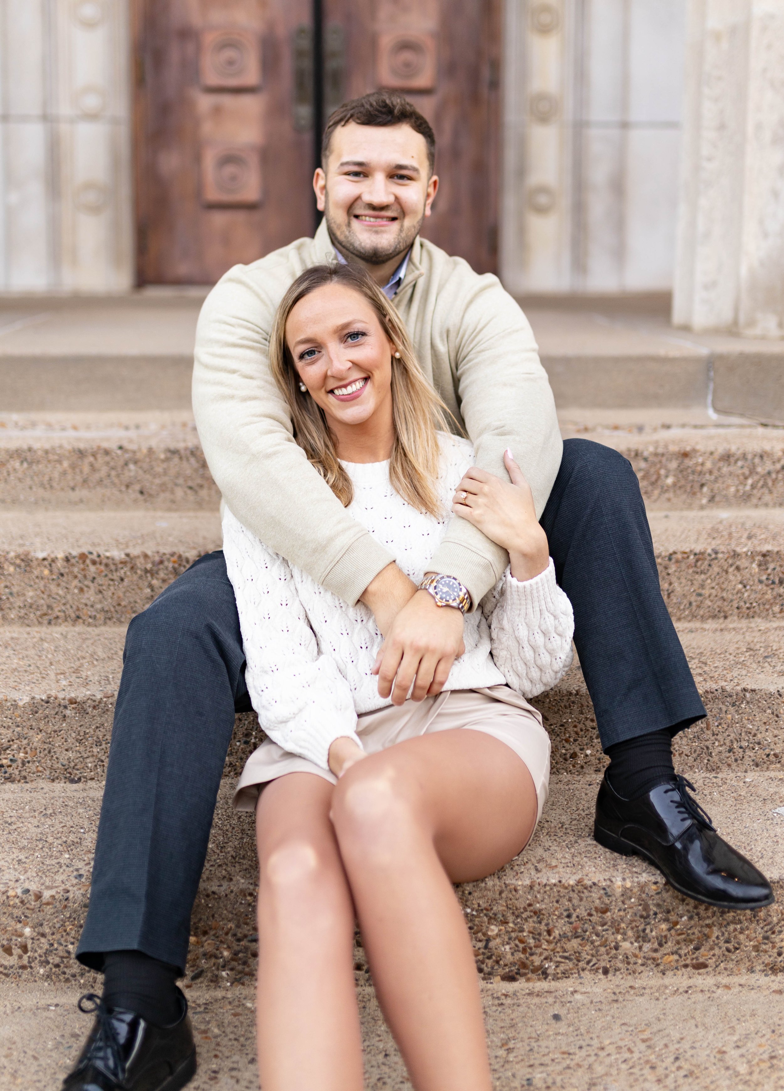 Young smiling couple sitting on outdoor steps, woman in a white sweater and tan shorts, man in a beige sweater and dark pants, in front of a large wooden door.