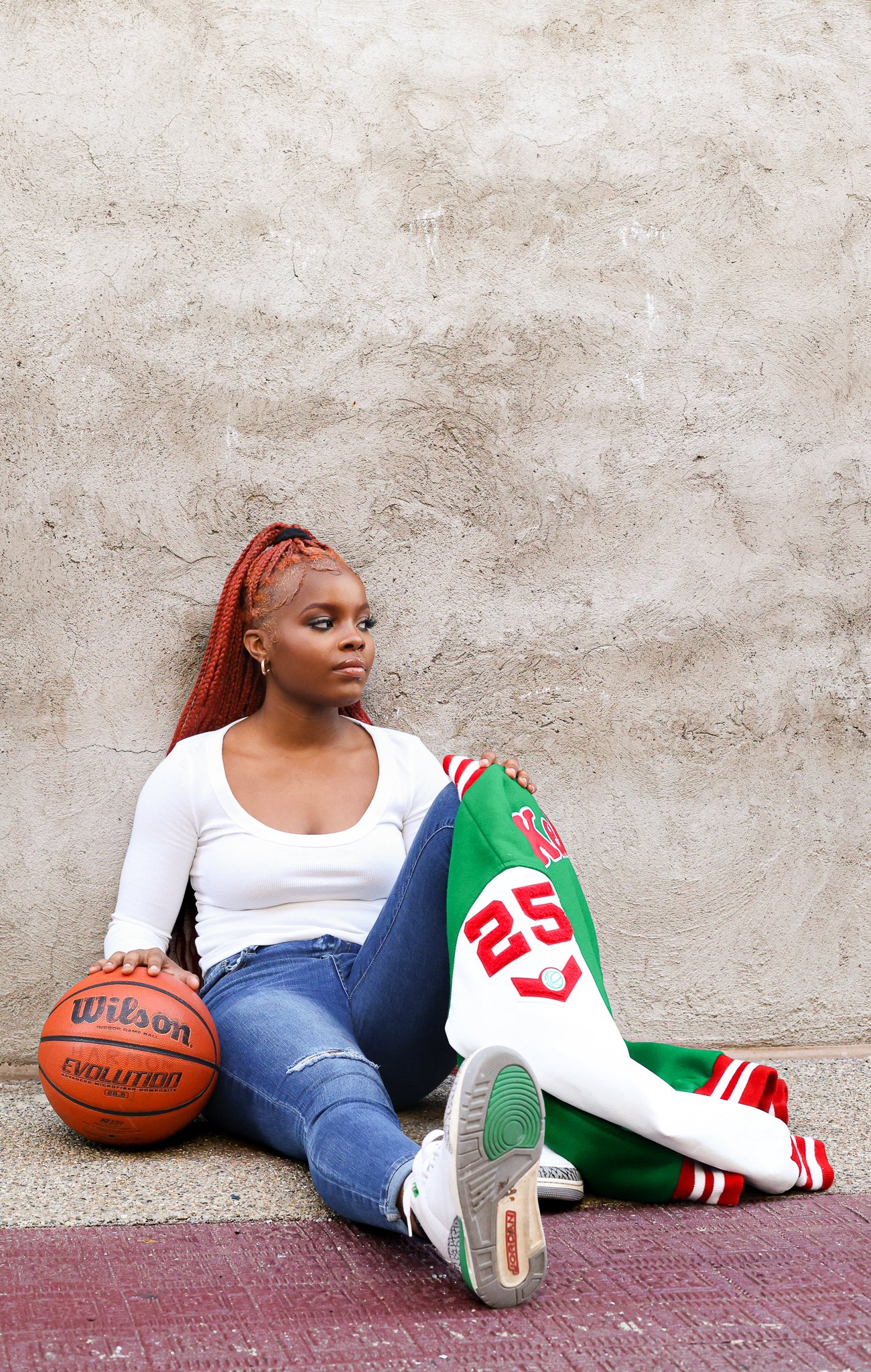 Young woman with long red braids sitting on the ground, holding a basketball, next to an Alabama Crimson Tide jacket, leaning against a beige wall.