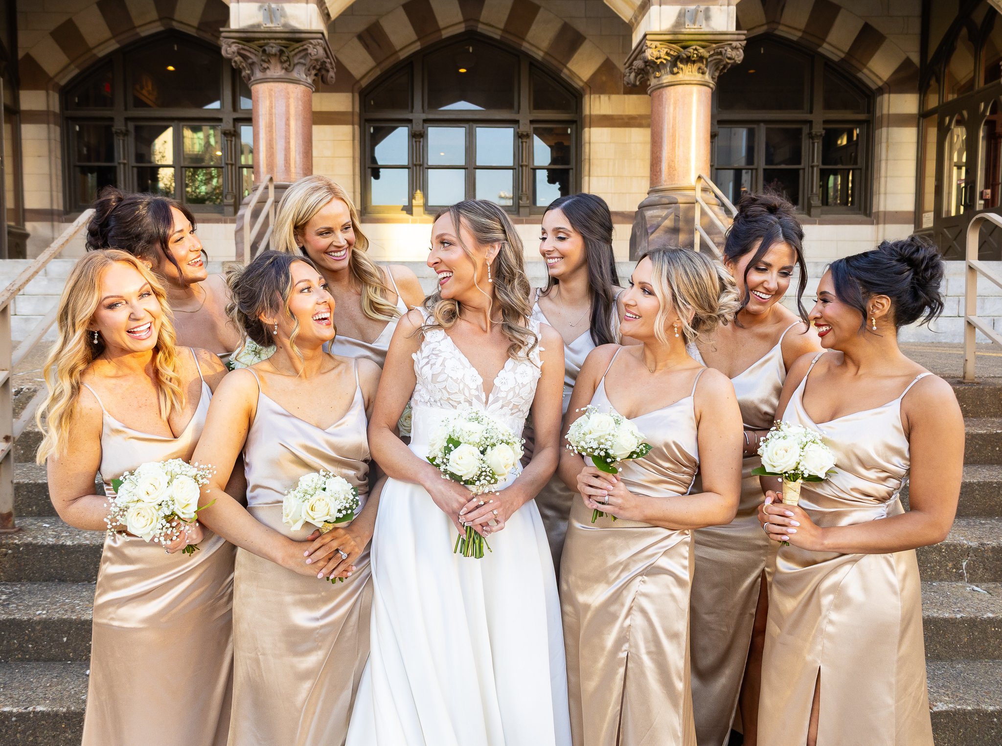 A bride and her bridesmaids standing on steps outside a building, all smiling and holding bouquets of white flowers.
