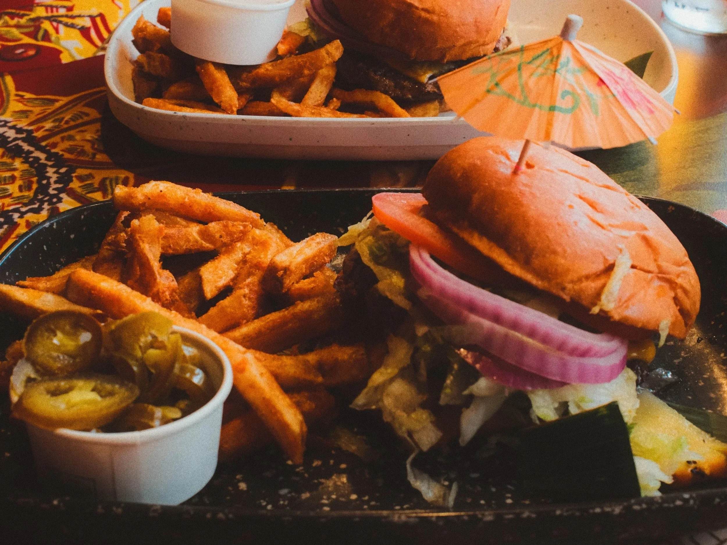 Close-up of a tray with a burger, French fries, and a small umbrella. A small cup of jalapeños is in the foreground. The background has vibrant, colorful tablecloths with patterns.