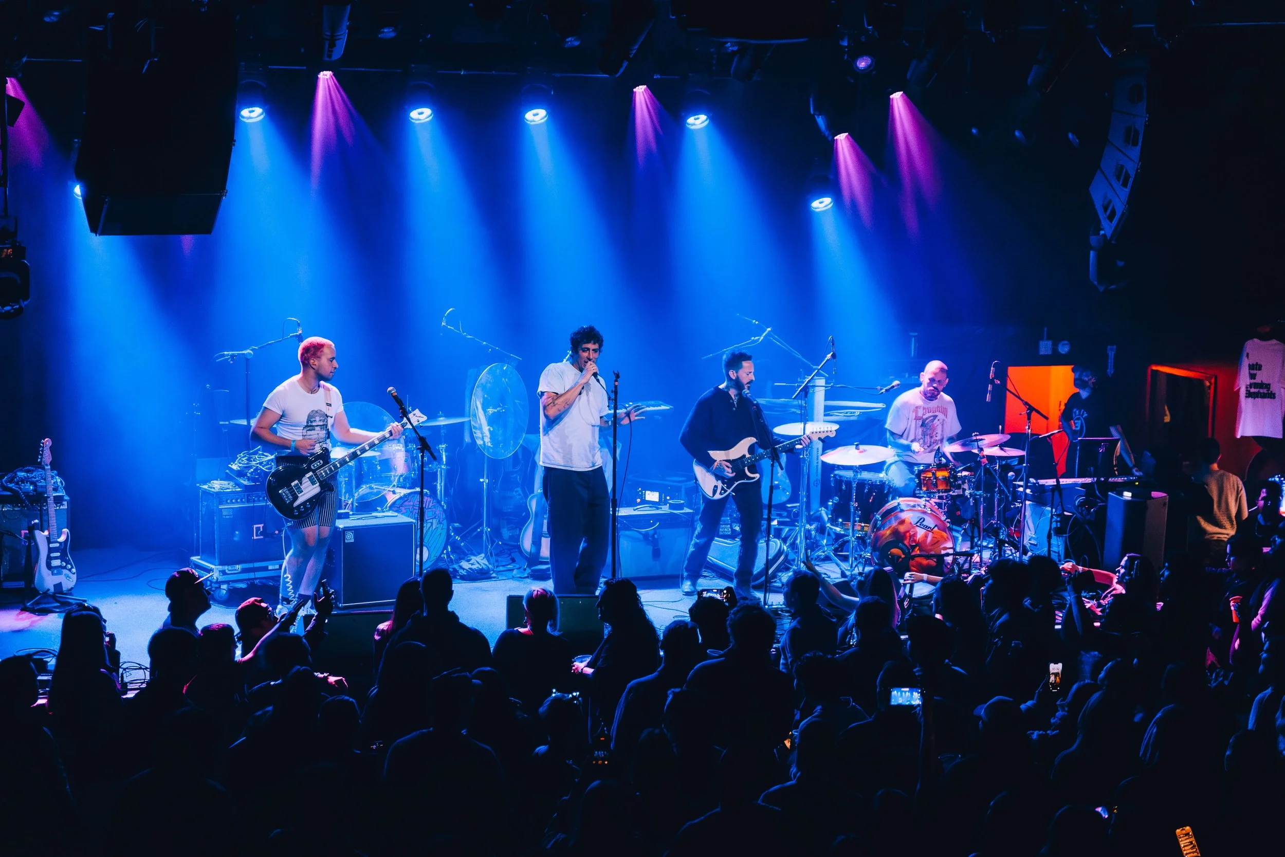 A live band performs on stage with various instruments including guitars, drums, and keyboards, lit by colorful stage lights, with an audience watching in the foreground.