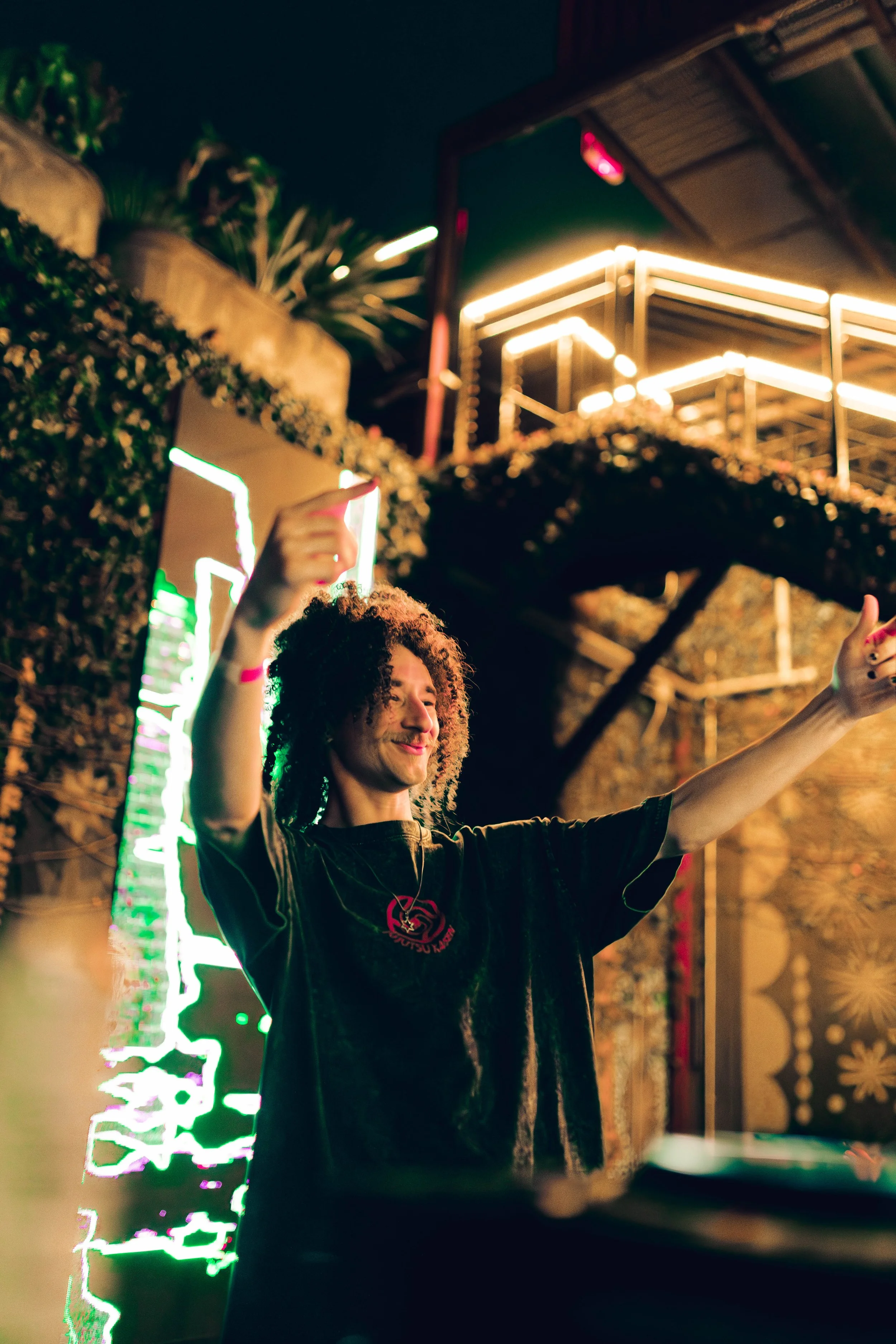 Young man with curly hair smiling and raising his hand at night in front of a decorated outdoor area with neon lights, plants, and wooden structures.