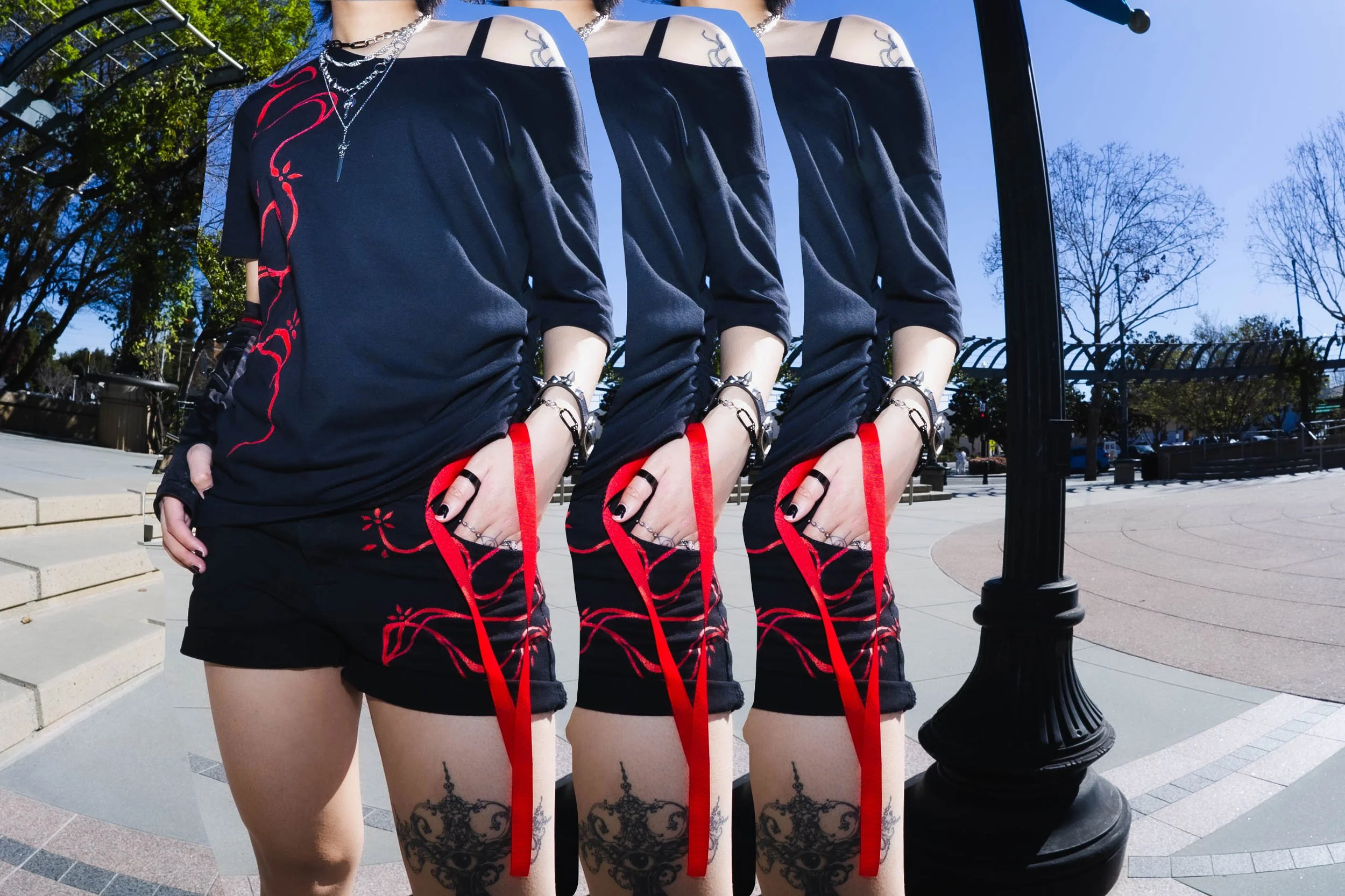 A woman wearing a black dress with red designs, black shorts, and various jewelry and tattoos on her legs, standing outdoors near a black lamppost with a reflective mirror behind her, creating multiple distorted reflections of her.
