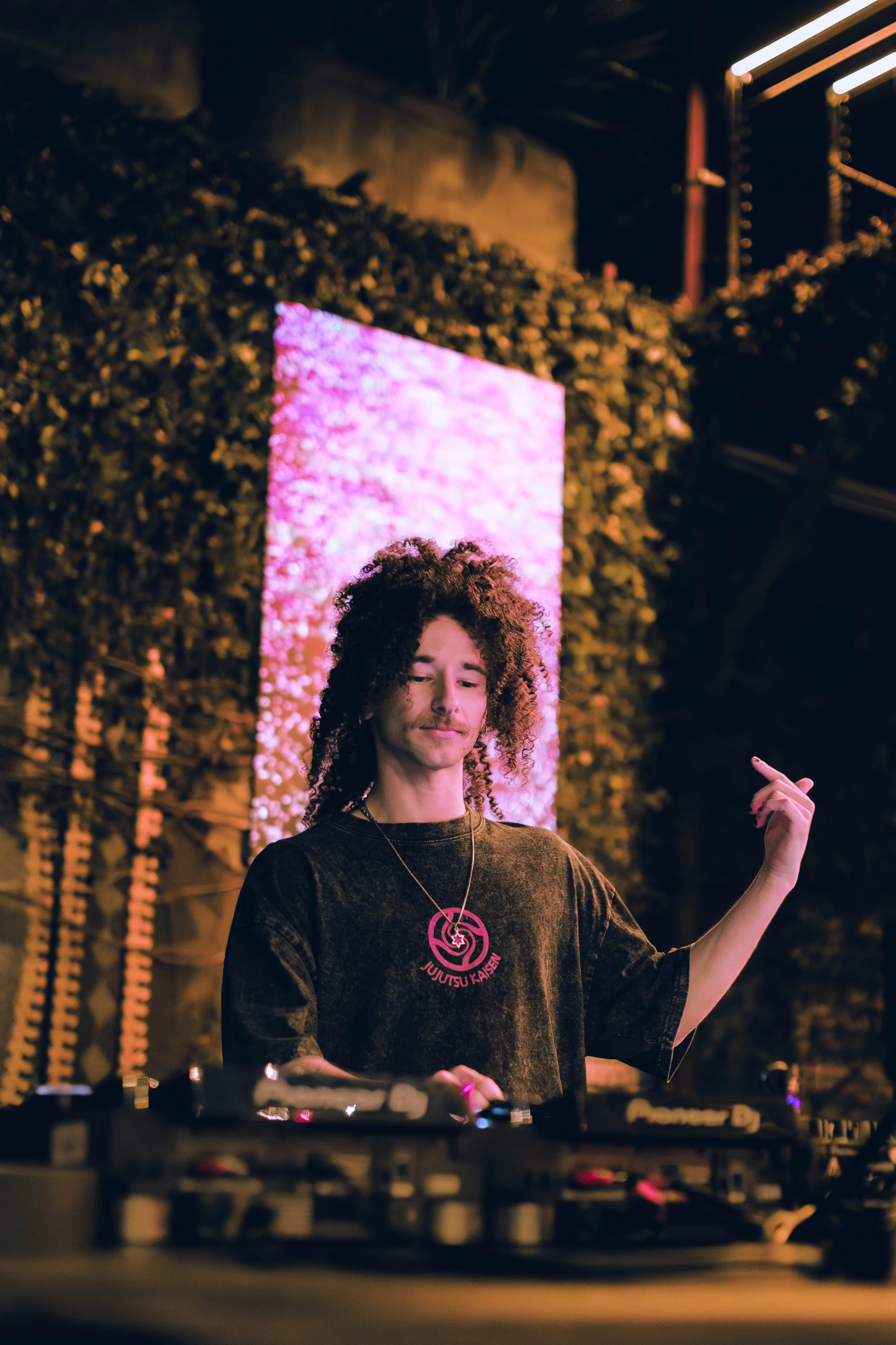 A DJ with curly hair wearing a black shirt with pink writing and a pink necklace, standing in front of a pink LED display and performing at an outdoor event at night.