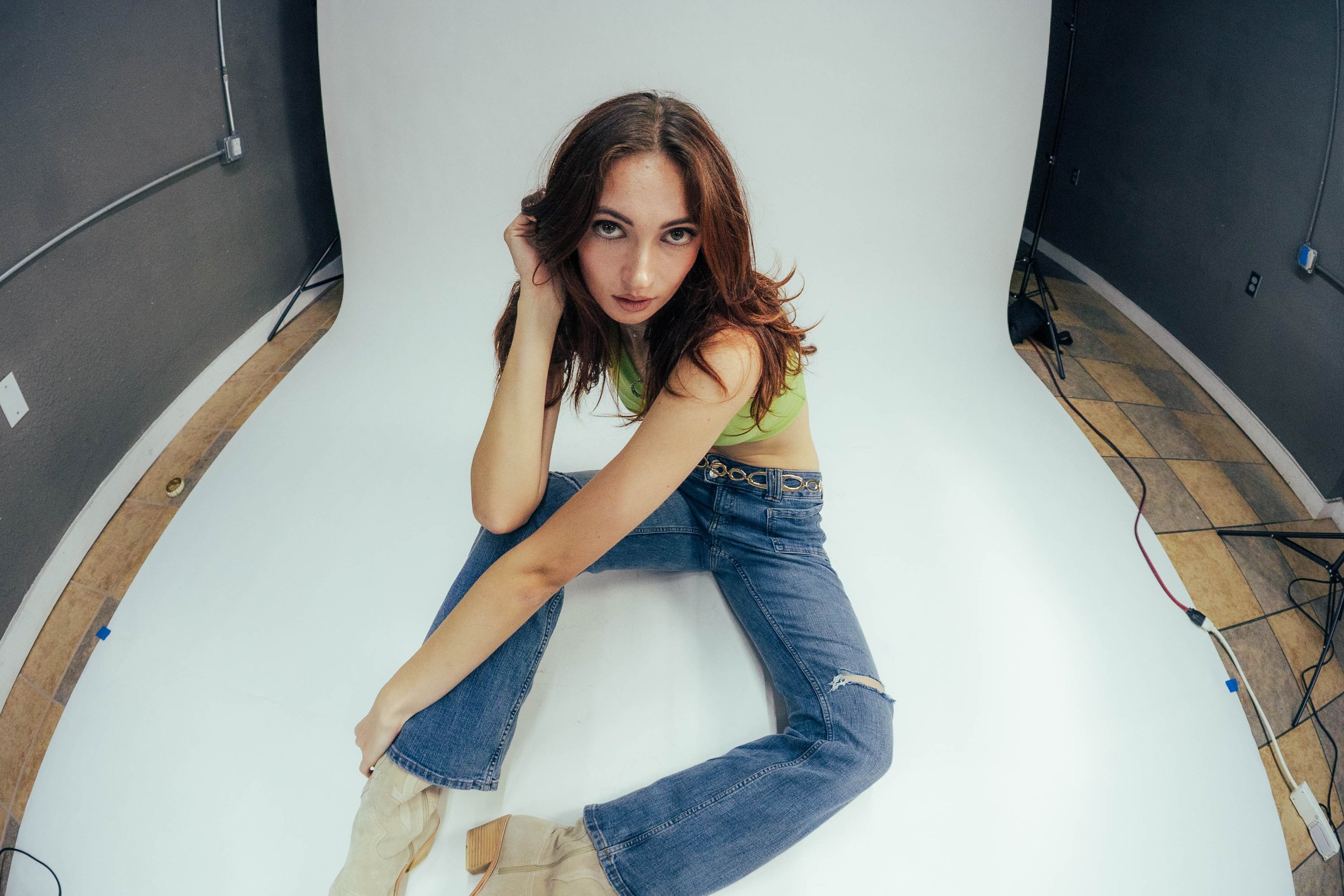 A woman with red hair and blue eyes sitting on her knees on a white photography backdrop, looking at the camera, wearing a green crop top, ripped jeans, and beige boots, in a photography studio with equipment and cords visible.