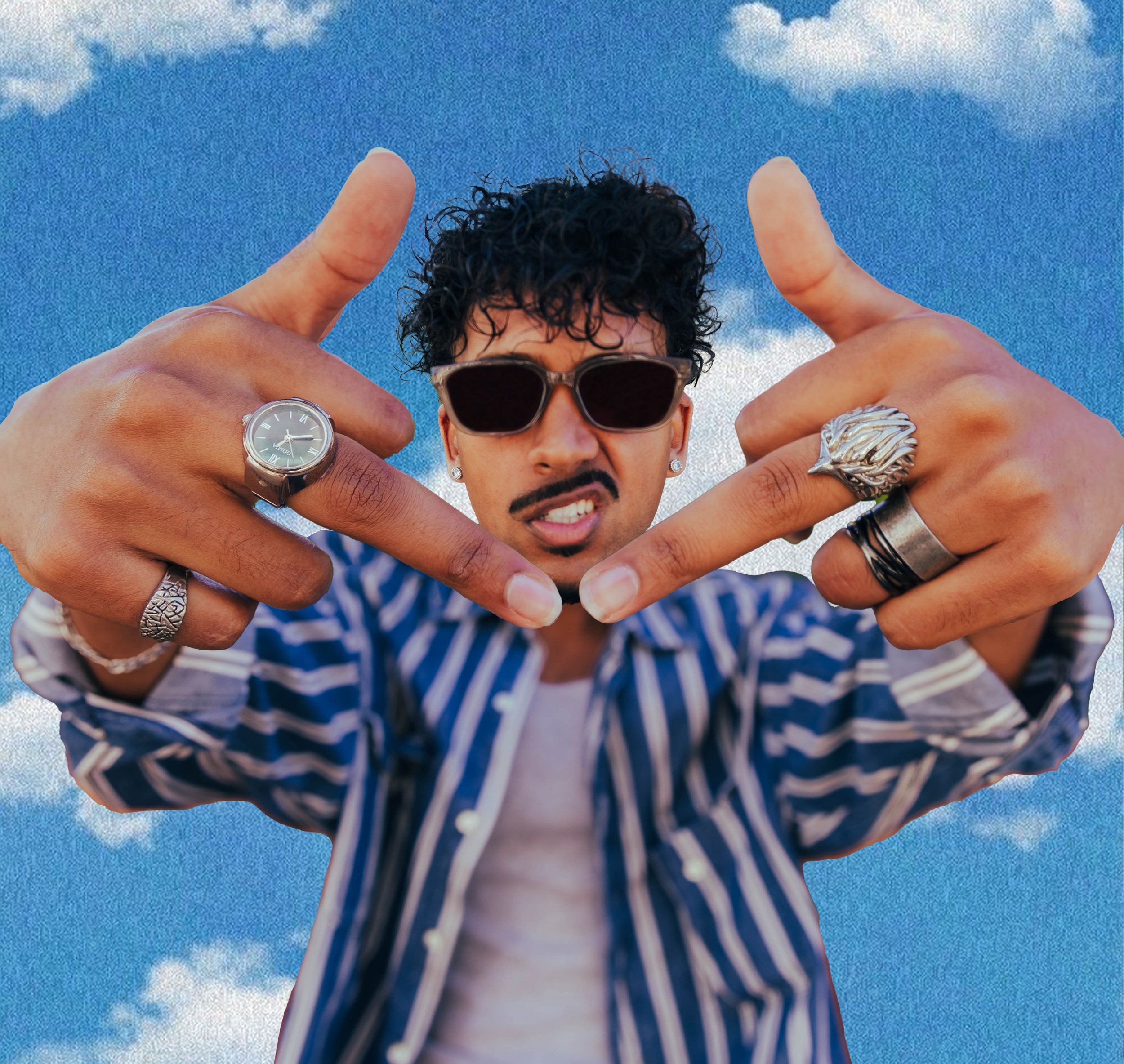 Person with dark curly hair wearing sunglasses, making a heart shape with their hands, wearing rings and a watch, against a background of blue sky and clouds.