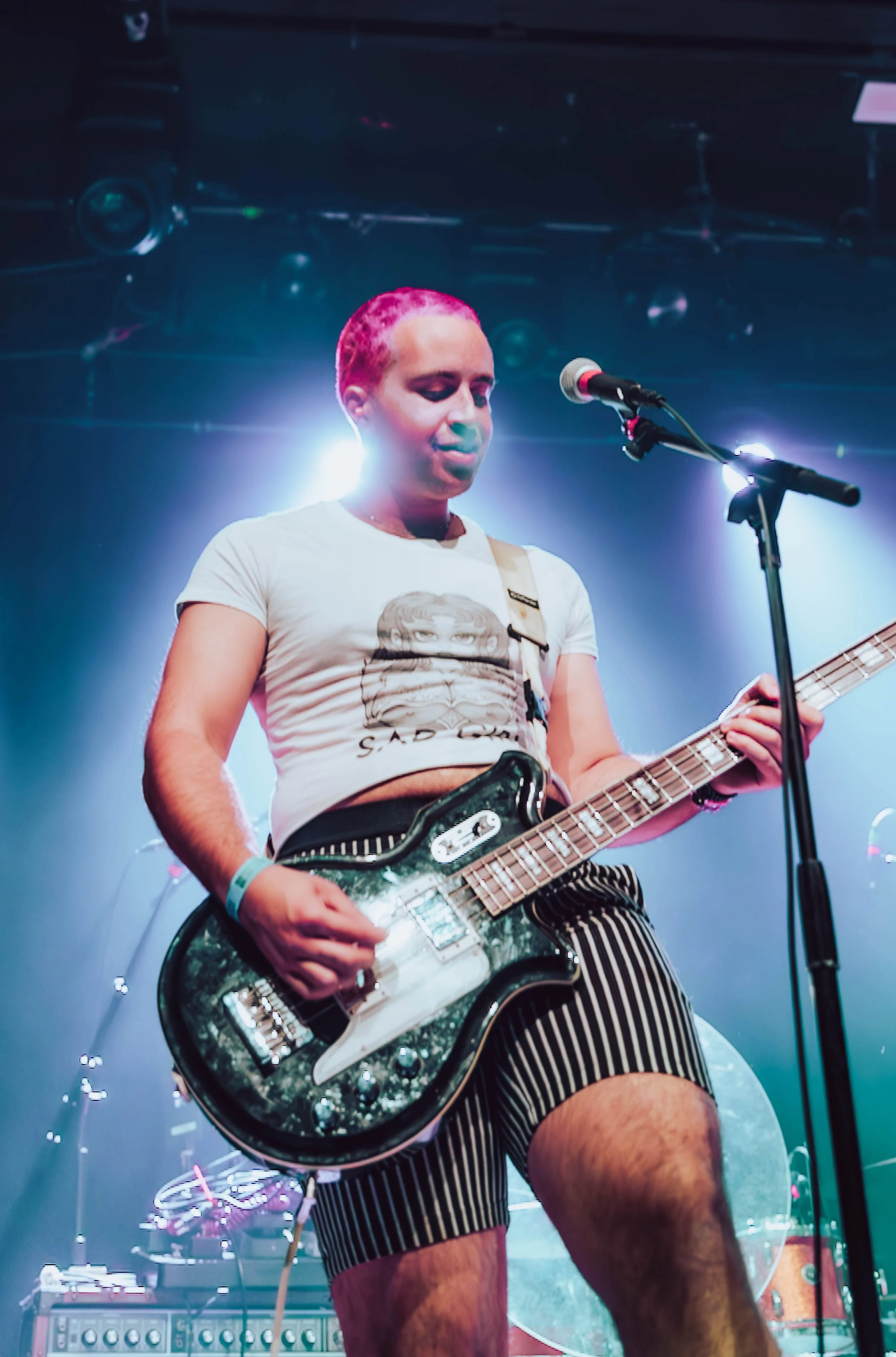 A person with pink hair playing an electric guitar on stage, illuminated by bright stage lights, with a microphone in front of them.