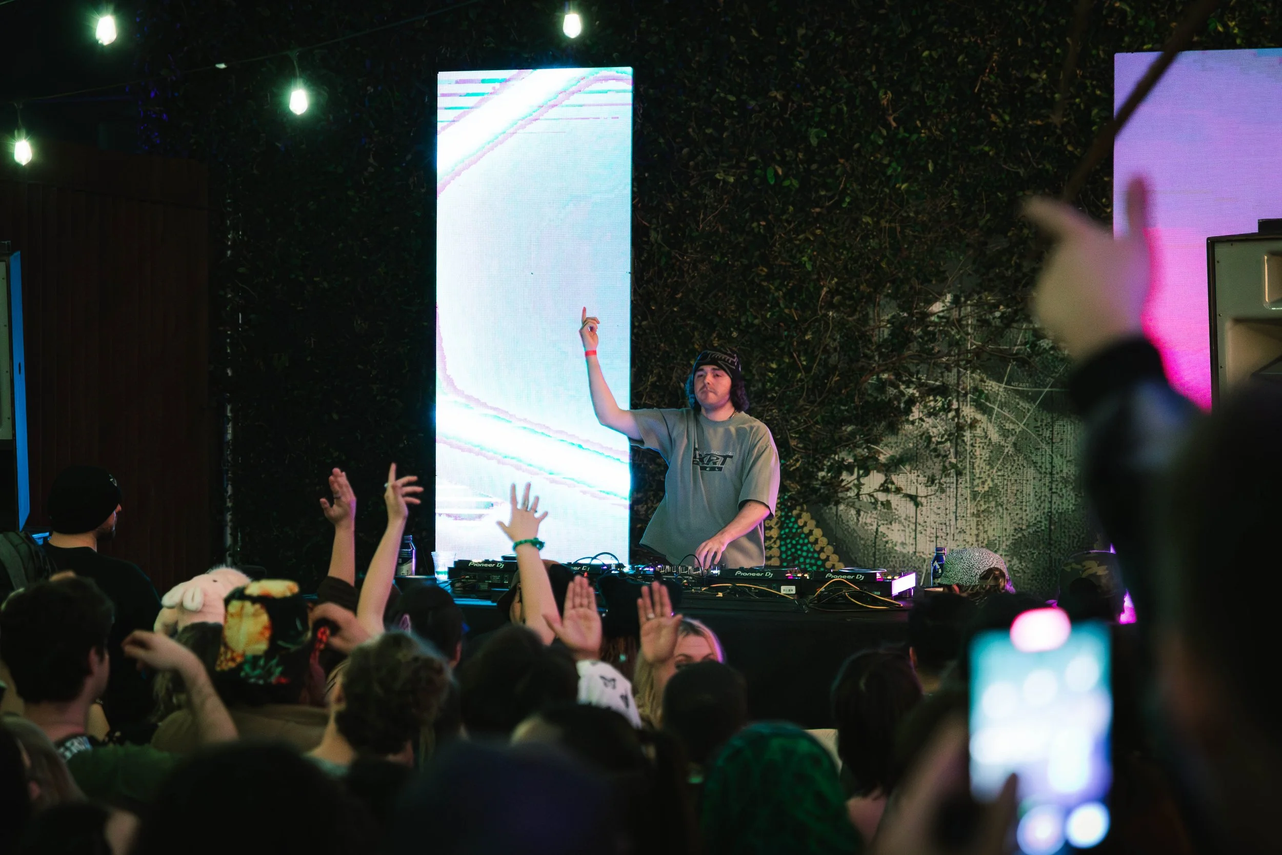 A DJ wearing a beanie and gray t-shirt is performing at an outdoor music event at night, with a crowd of people dancing and raising their hands. There are LED screens and string lights overhead.