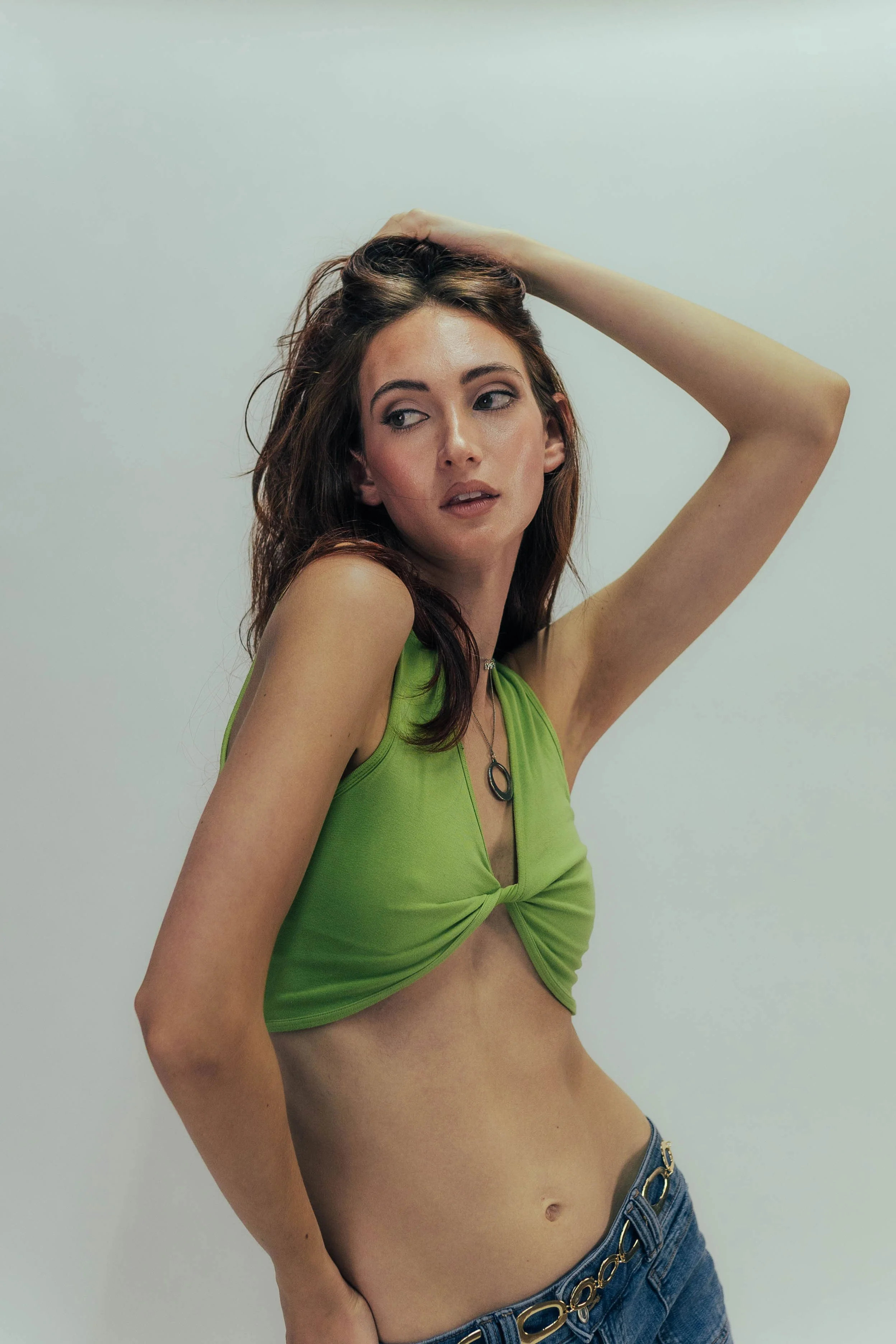 A young woman with brown hair, wearing a green cropped top and blue jeans, posing with one hand on her head against a plain white background.