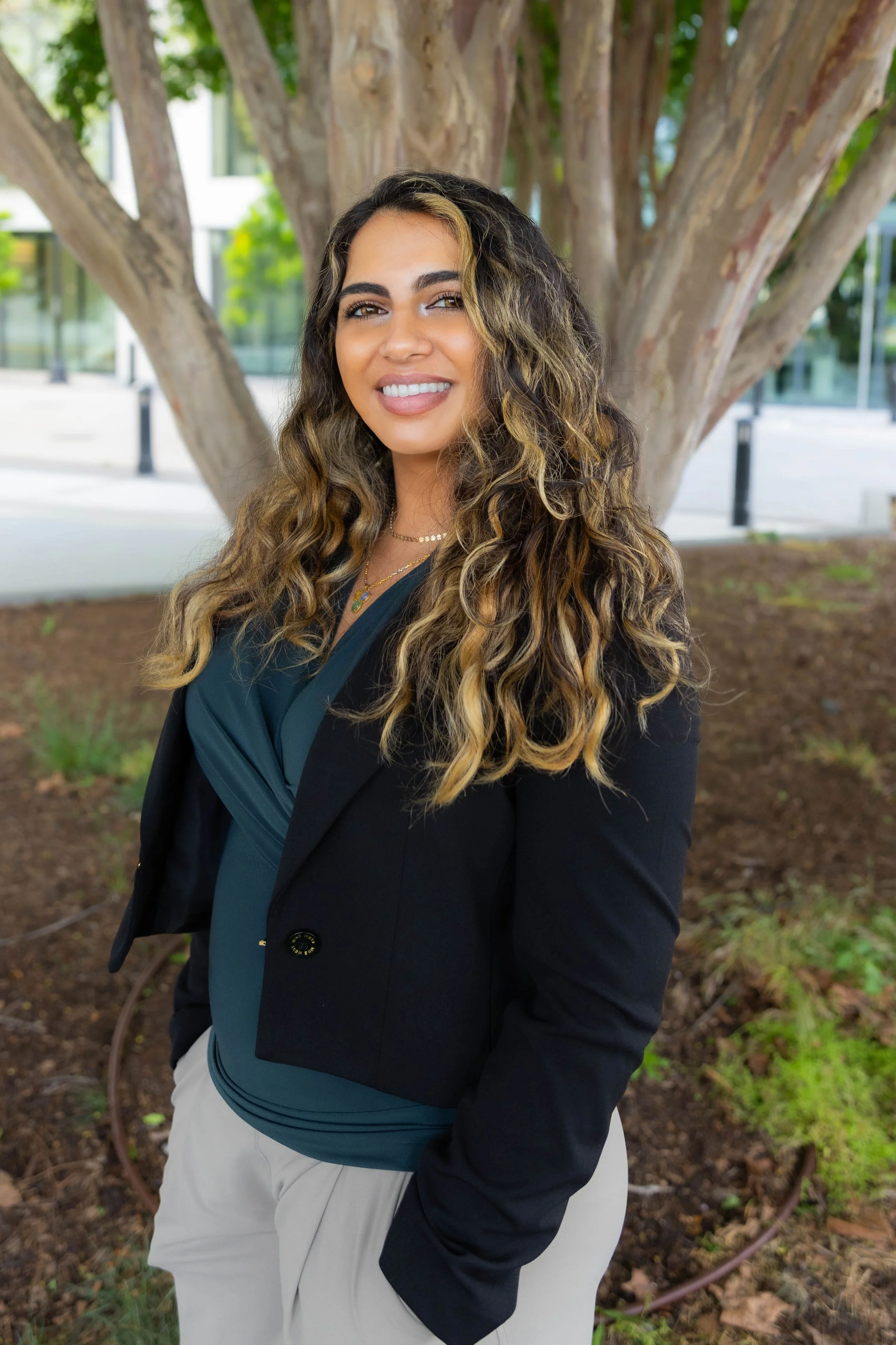 A woman with long, curly hair blonde and dark, smiling, standing outdoors in front of a tree with a modern building in the background.