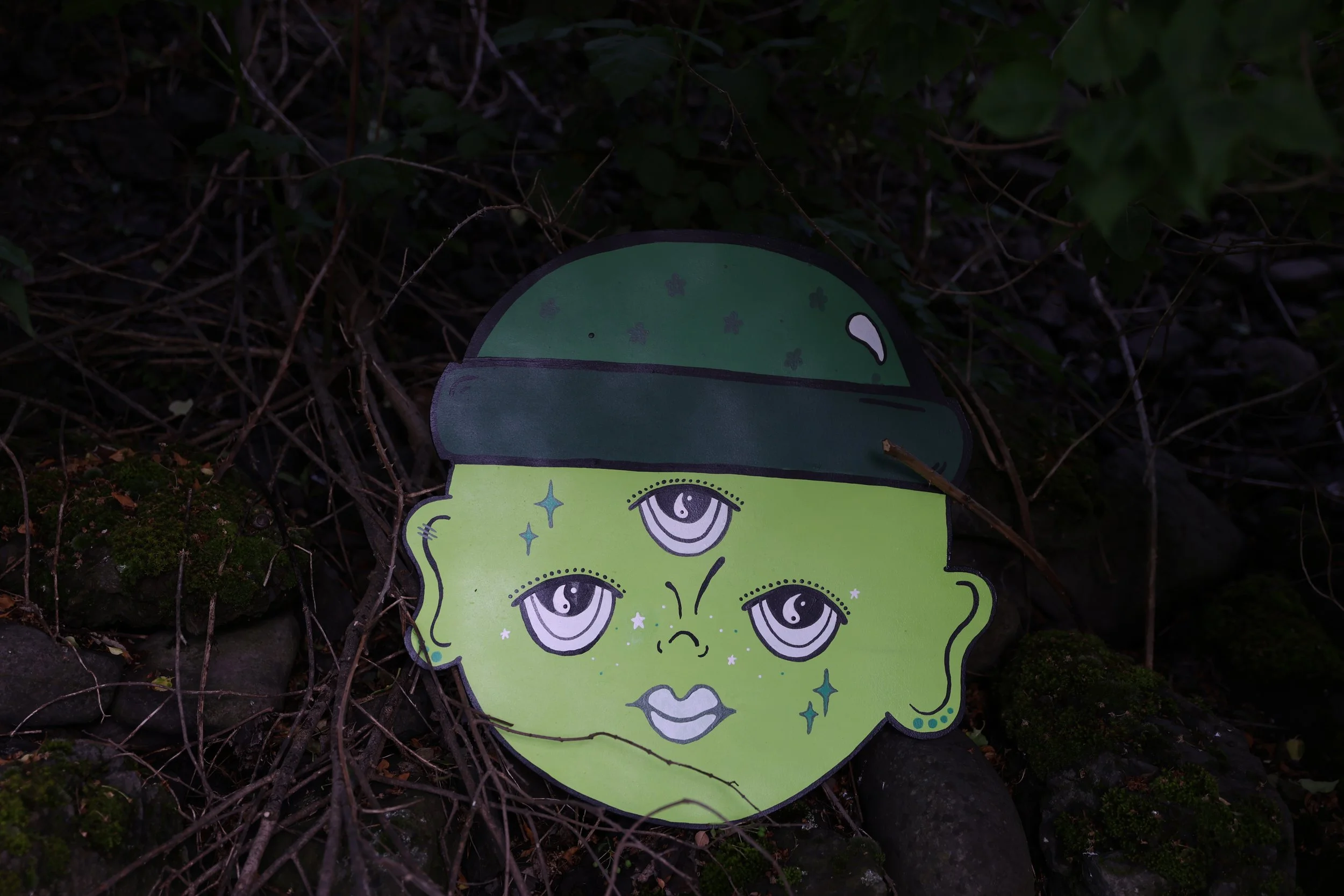 A cartoonish green face with multiple eyes and lips, surrounded by small stars, partially hidden among dark foliage and twigs on the ground.