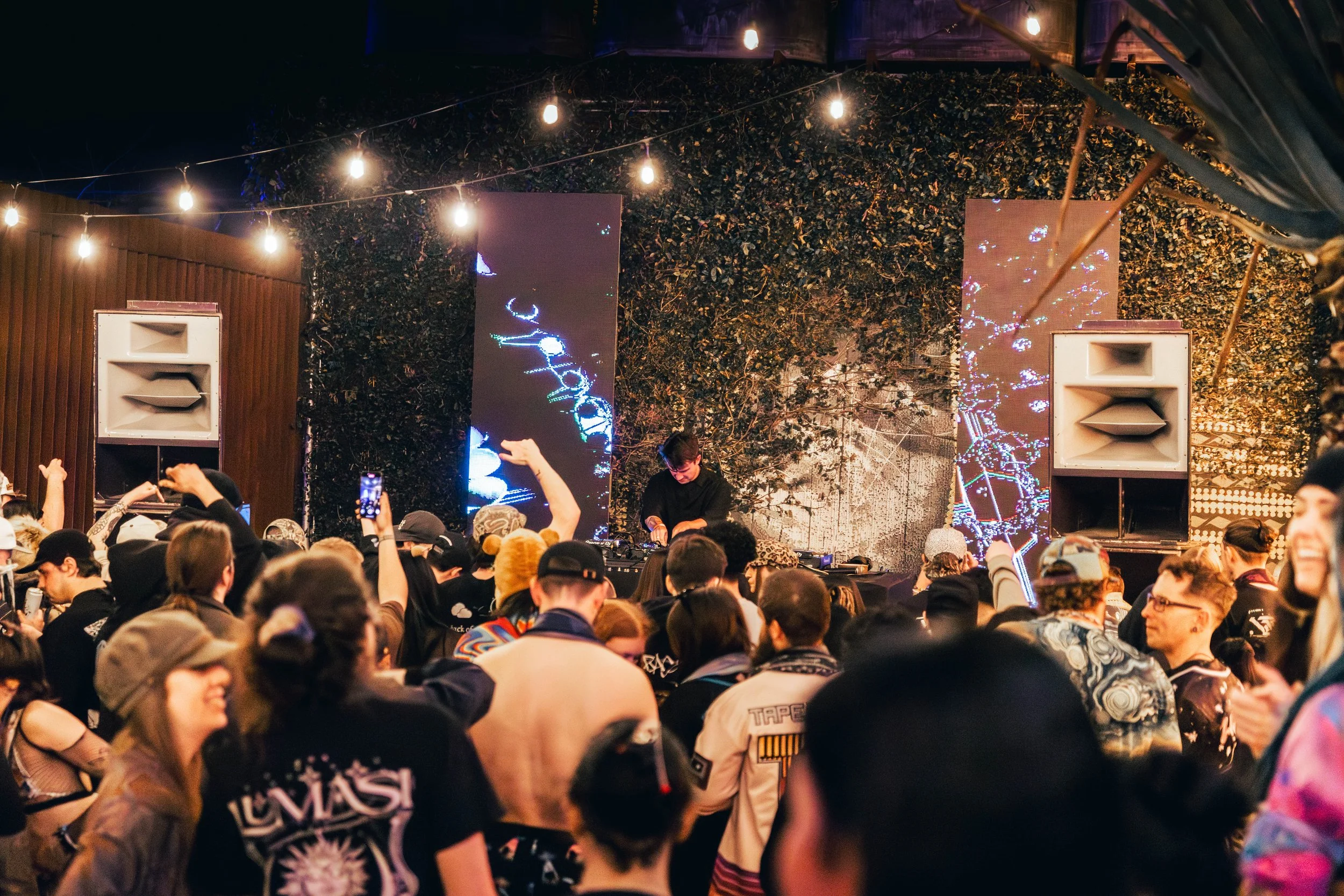 A crowd of people at a music event with a DJ performing in front of large screens and string lights overhead, set against a backdrop of greenery.
