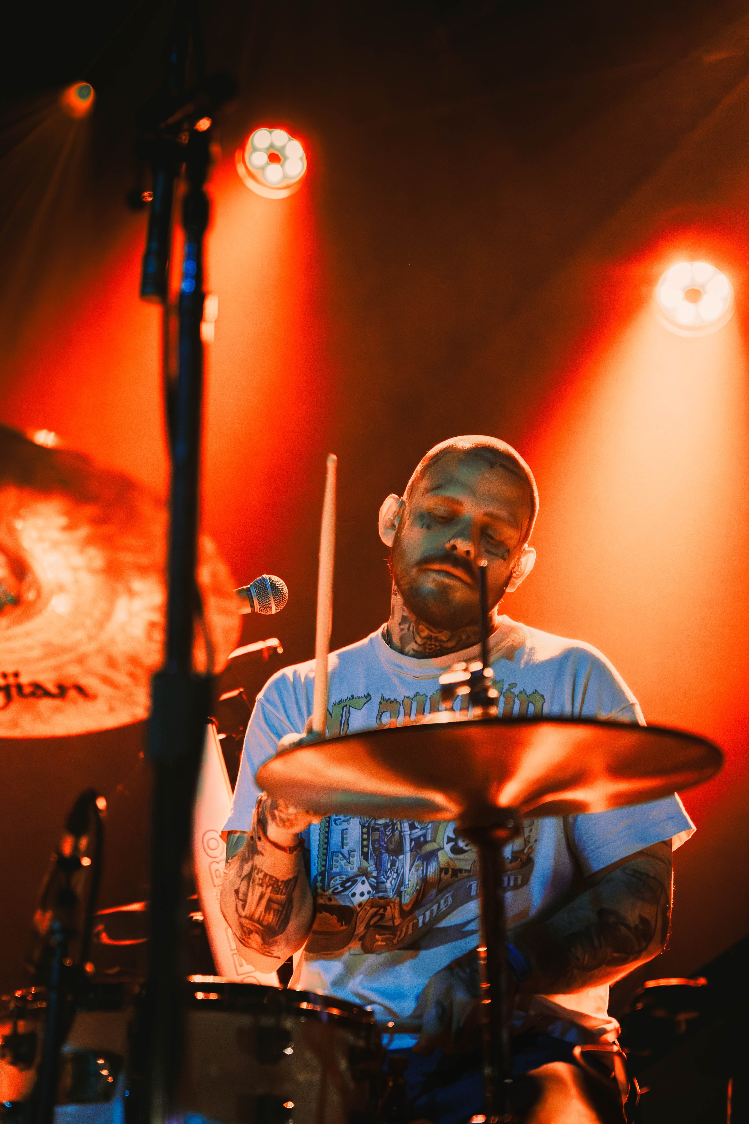 A male drummer with tattoos on his arms and neck is performing on stage. The stage is lit with warm orange and red lights, and he is focused on playing his drum set, with various drum sticks and microphones visible.