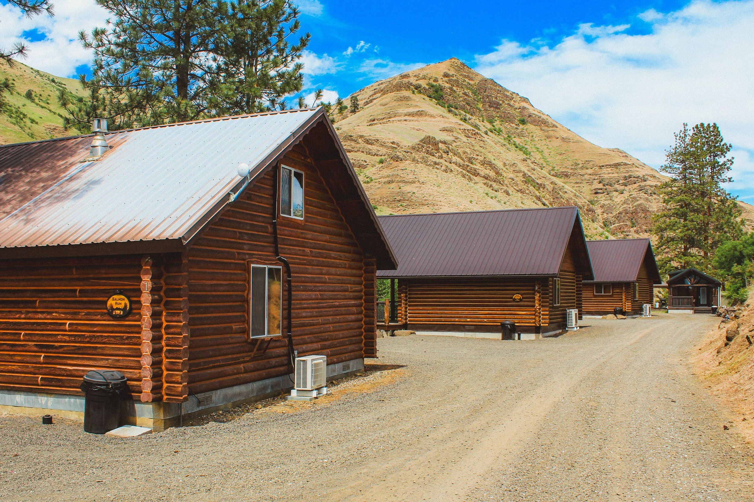 A row of log cabins with metal roofs in a mountainous area, trees, and a gravel ground under a partly cloudy sky.