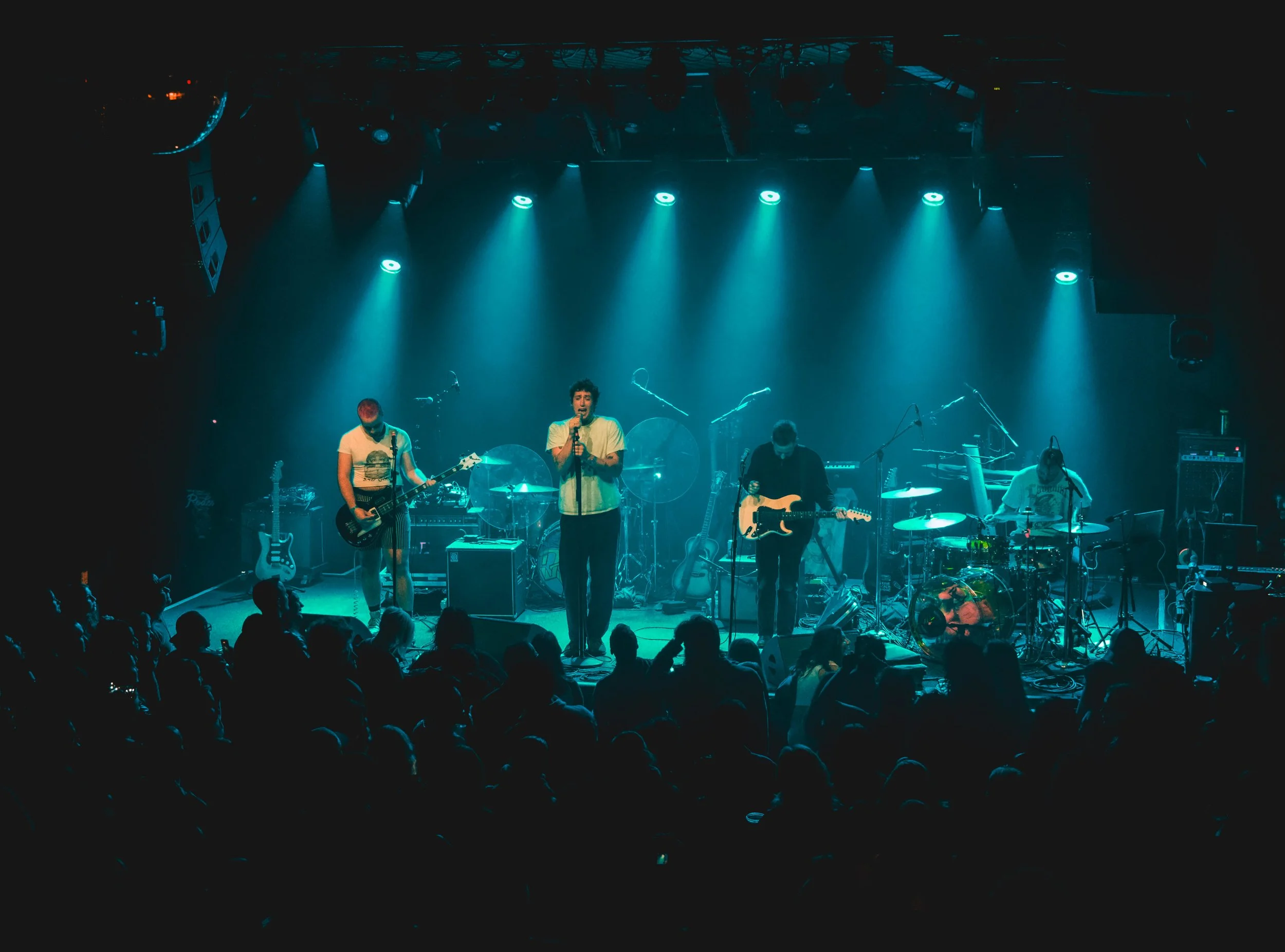 A band performs on stage under blue and white spotlights with audience in front.