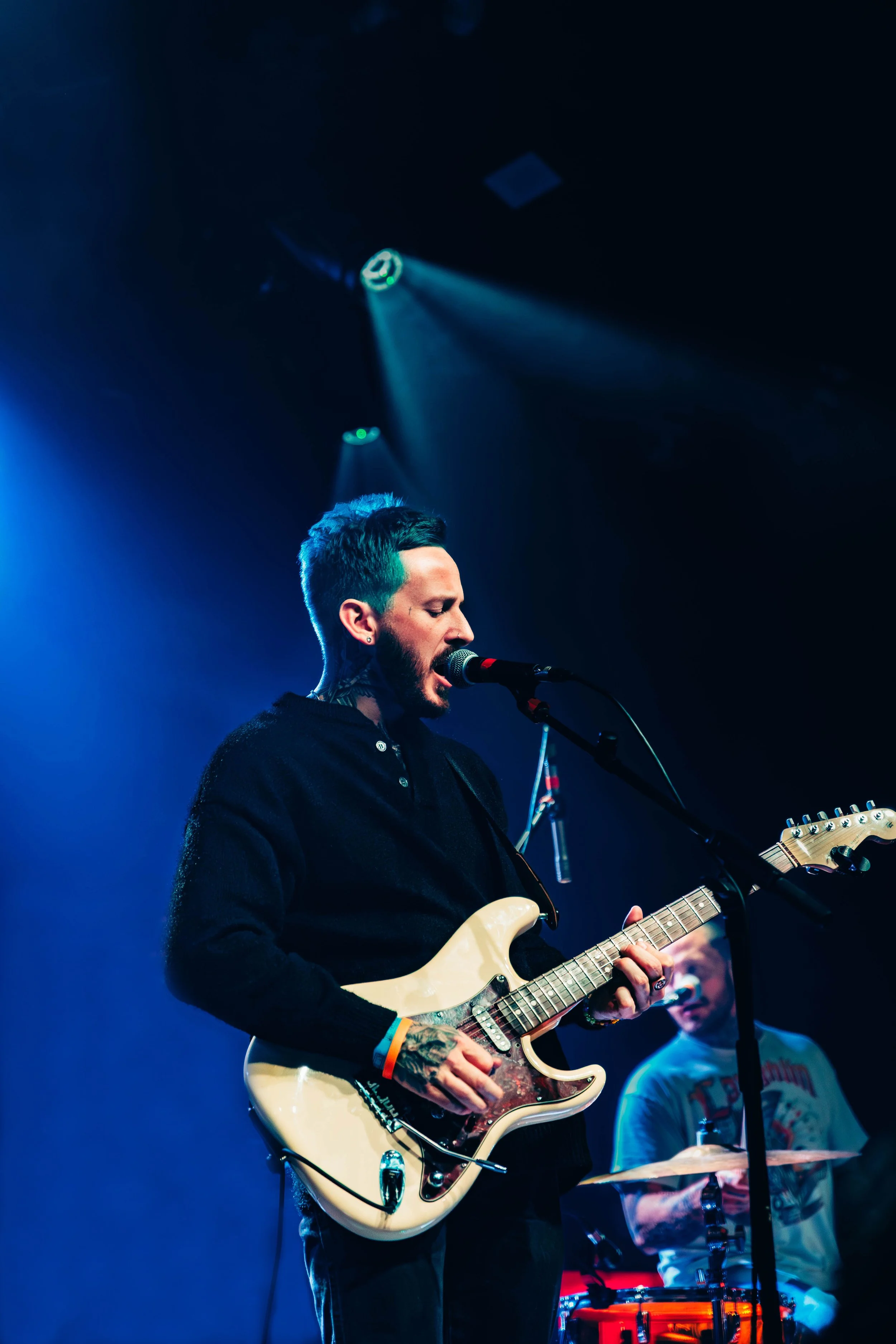 A male musician with tattoos wearing a black jacket, singing into a microphone, and playing an electric guitar on stage with blue and purple lighting, while another band member is visible in the background.