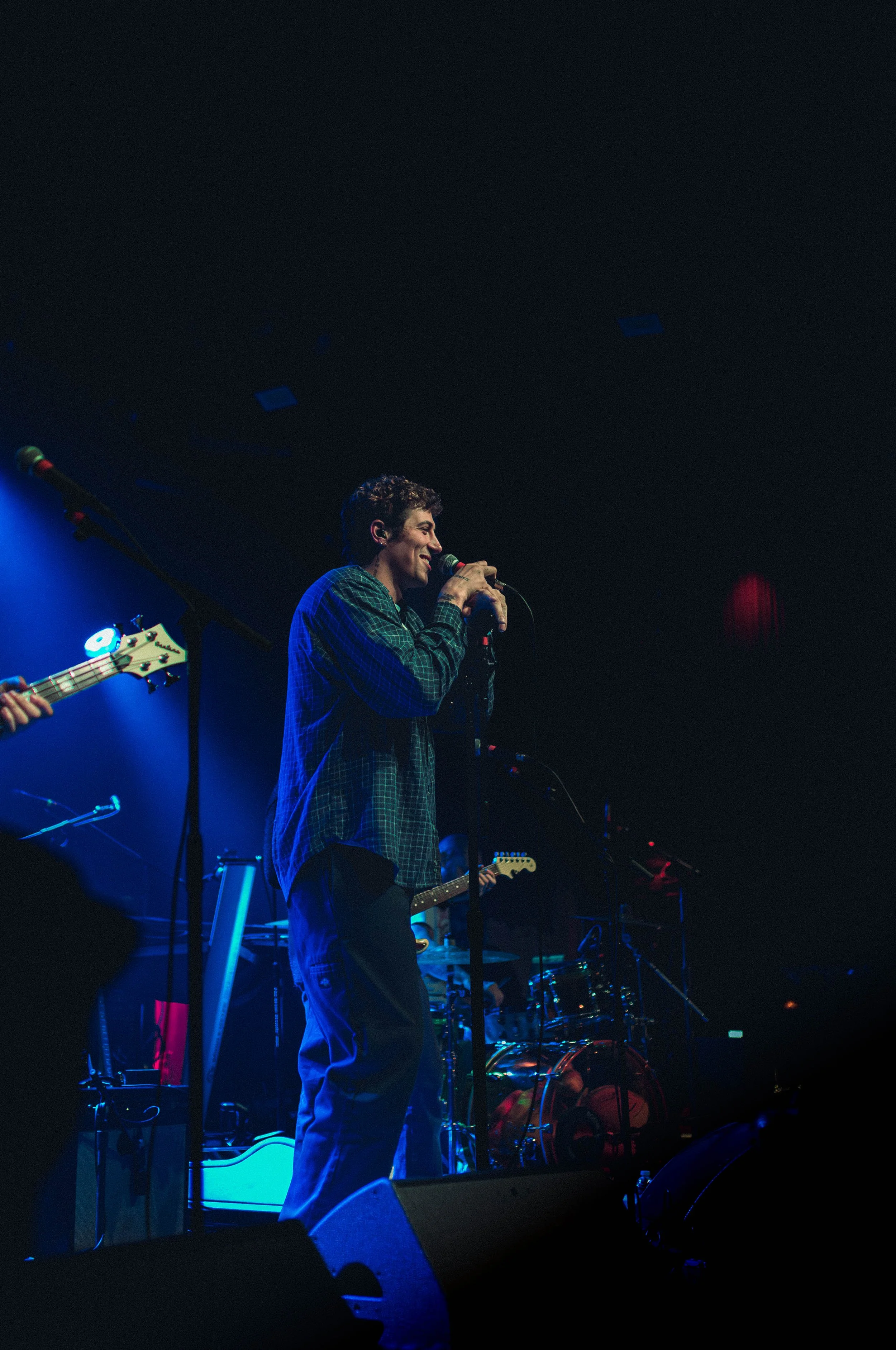A man singing into a microphone on a stage with musical equipment surrounding him, illuminated by blue stage lights.