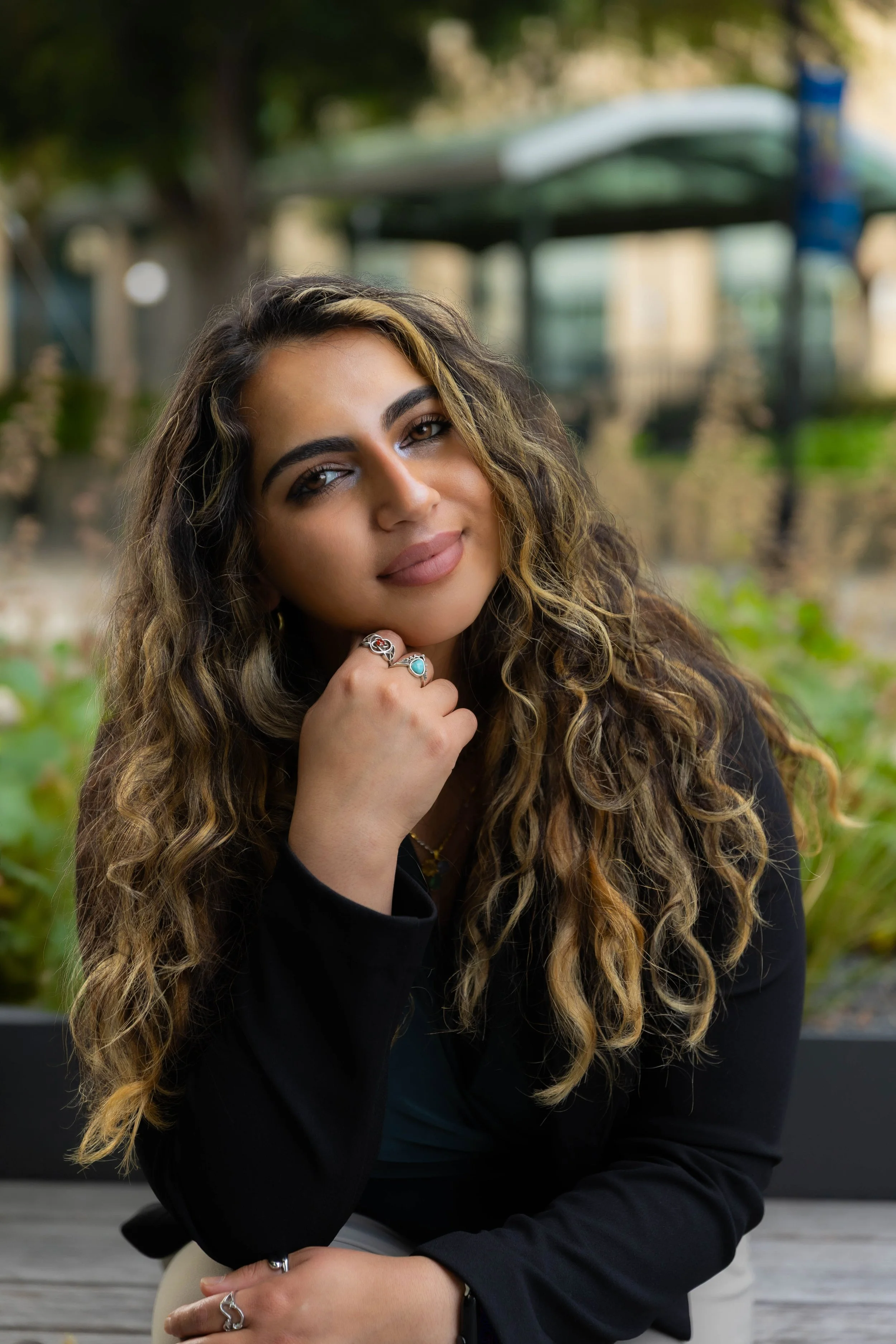 A young woman with long curly hair and hoop earrings, smiling softly with her chin resting on her hand, sitting outdoors in a park-like setting with blurred trees and a covered structure in the background.