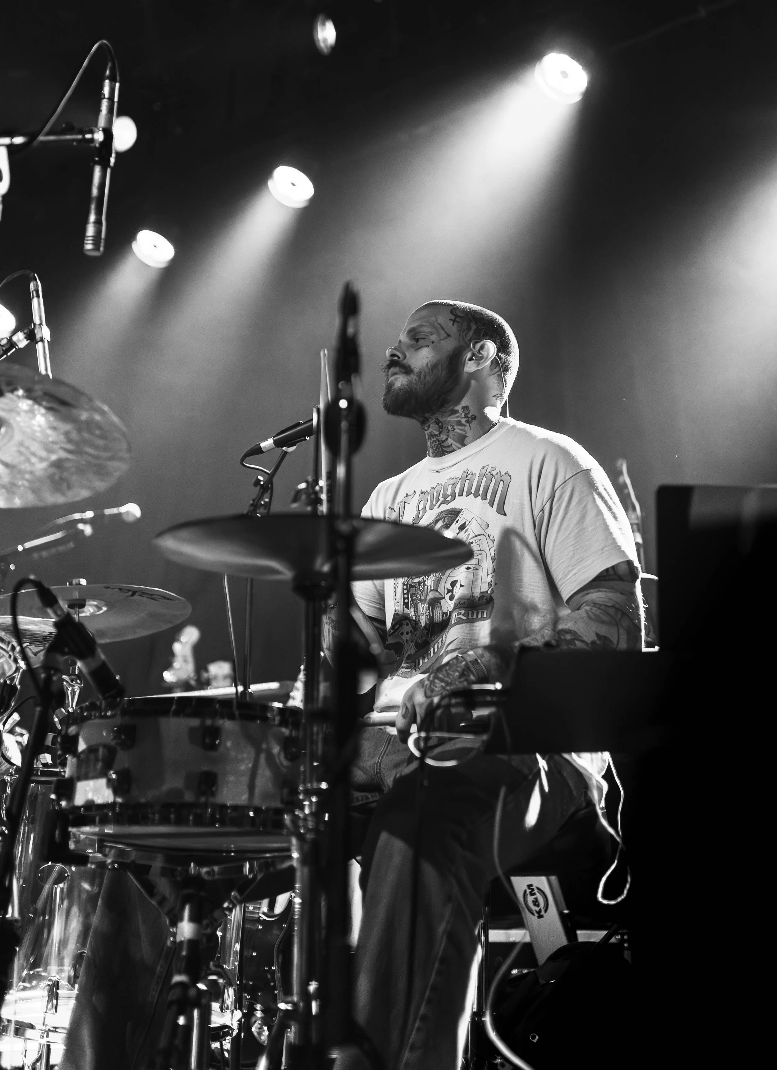 A musician with tattoos and a beard playing the drums on stage, with stage lights shining down in the background.