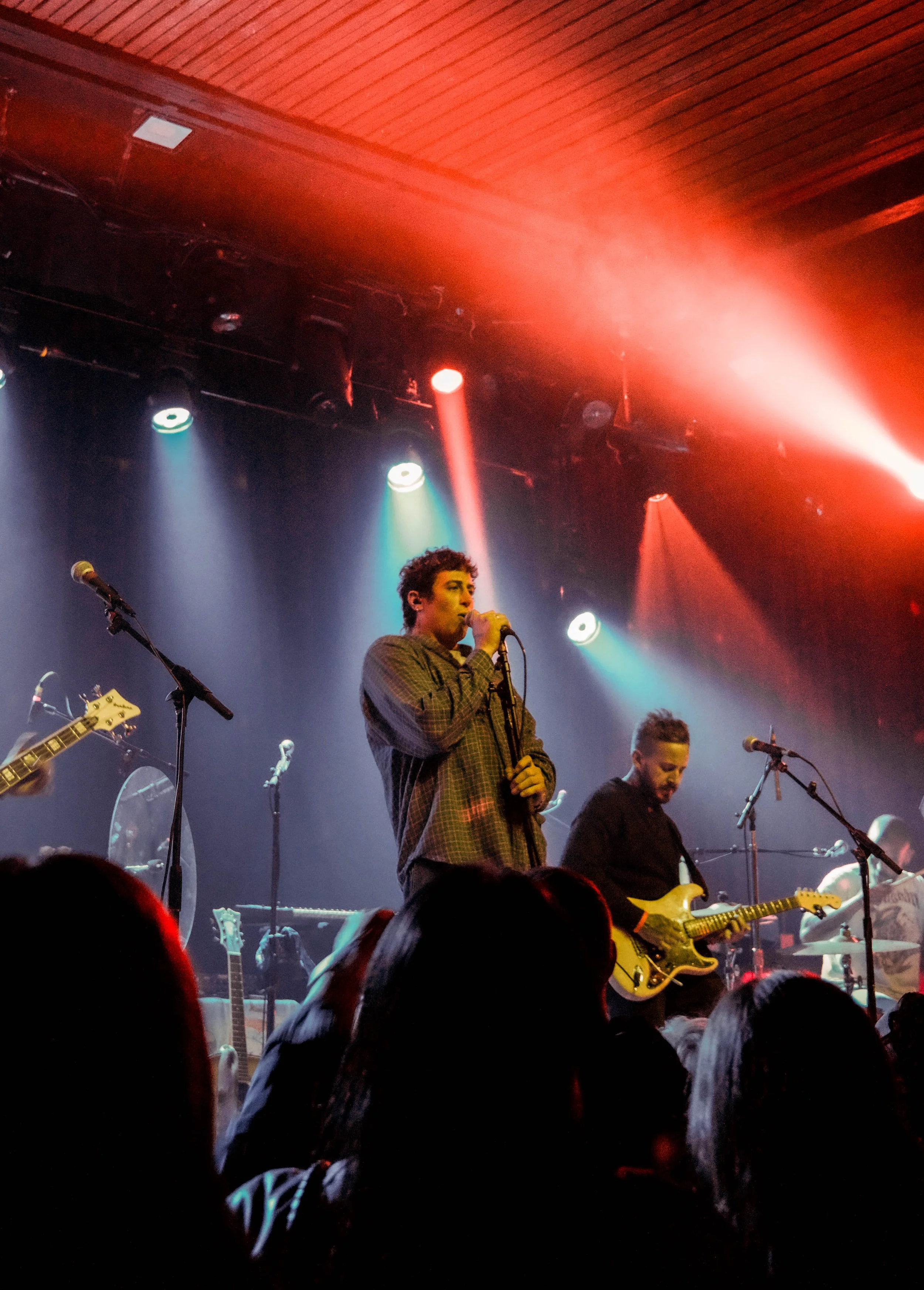 A band performs on stage in a dimly lit venue with colorful stage lights and a wooden ceiling, featuring a singer holding a microphone and a guitarist playing an electric guitar.