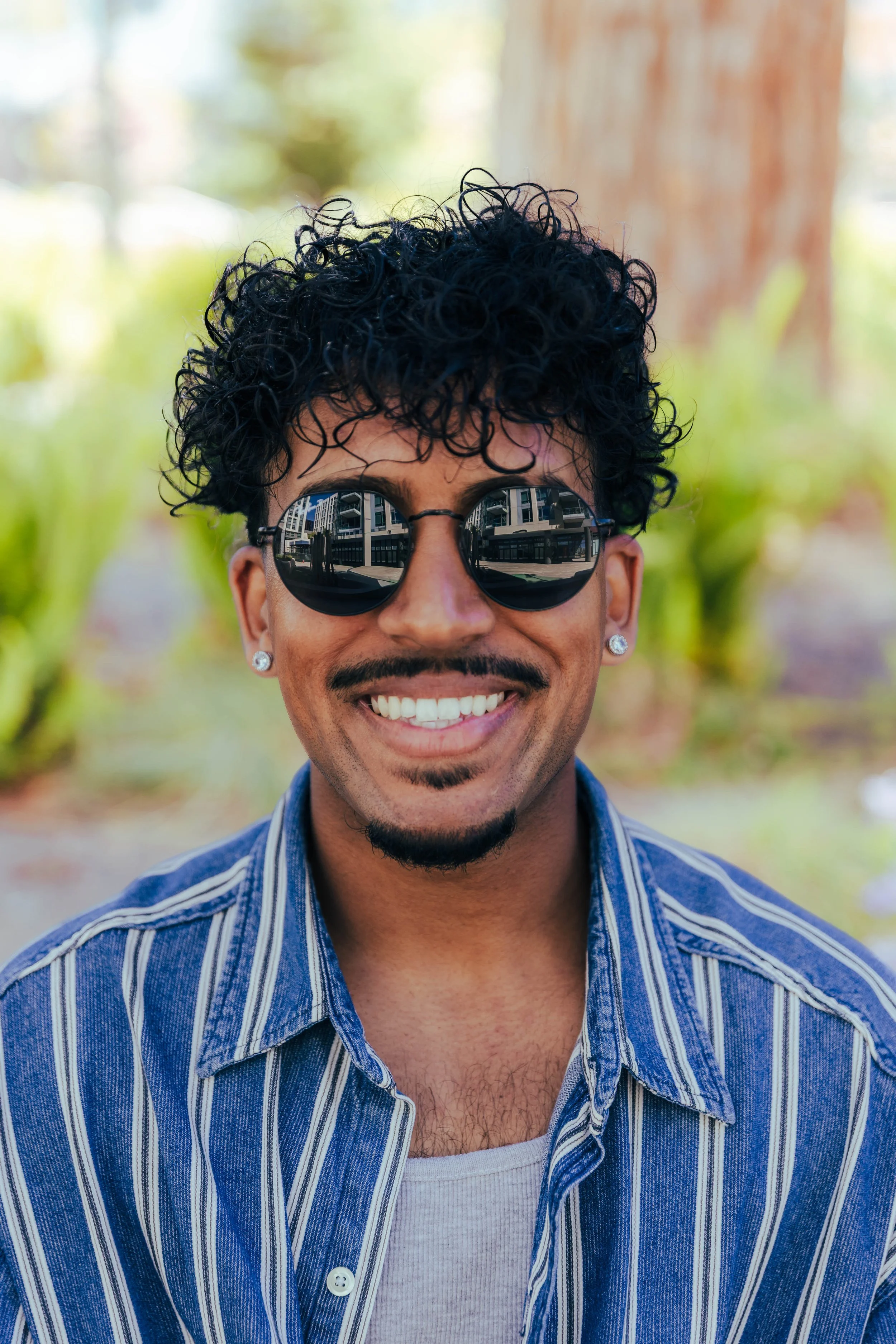A man wearing reflective sunglasses with a cityscape reflected in the lenses, smiling outdoors with trees and a wooden structure in the background, dressed in a striped blue and white shirt over a white t-shirt.