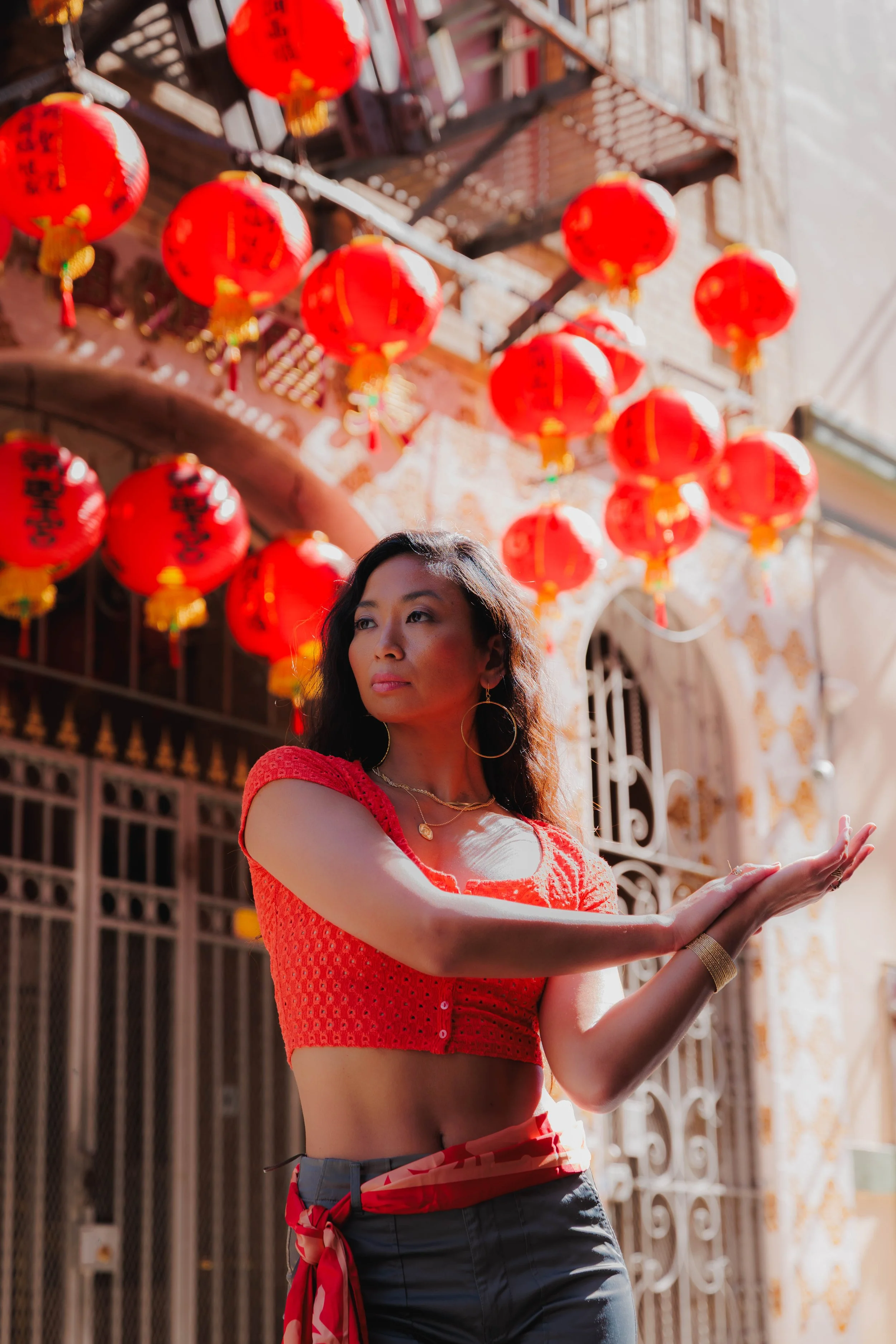 Woman wearing a red textured crop top, gold jewelry, and dark pants, standing with her arms extended in front of hanging red lanterns on a street with ornate buildings.