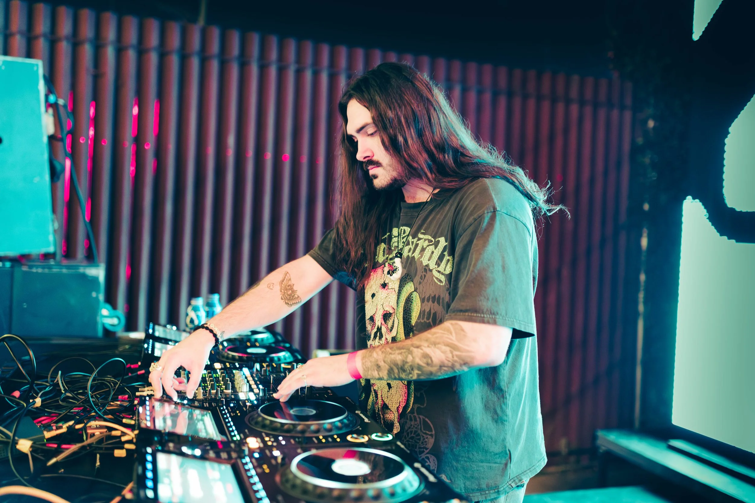 A male DJ with long hair and tattoos DJing with equipment in a dark, colorful venue.