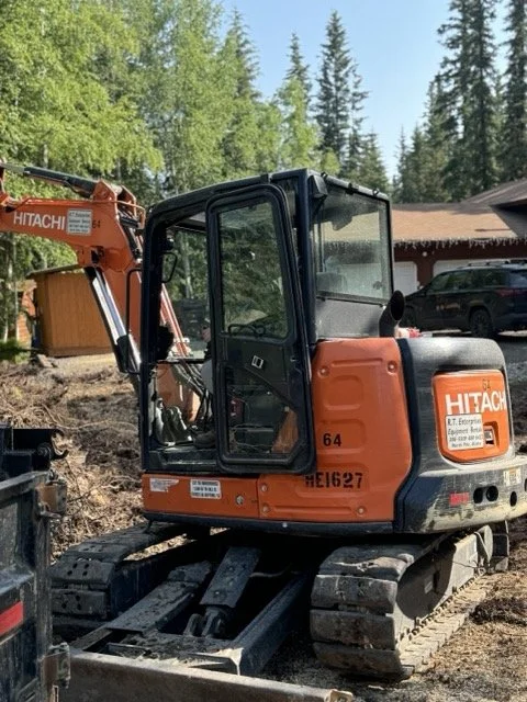 Orange Hitachi mini excavator parked on dirt yard with trees and house in background.