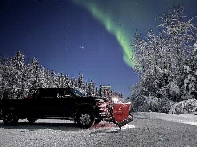 A black pickup truck with a red snowplow attached to the front parked on a snowy road during night, with snow-covered trees and the Northern Lights visible in the sky.