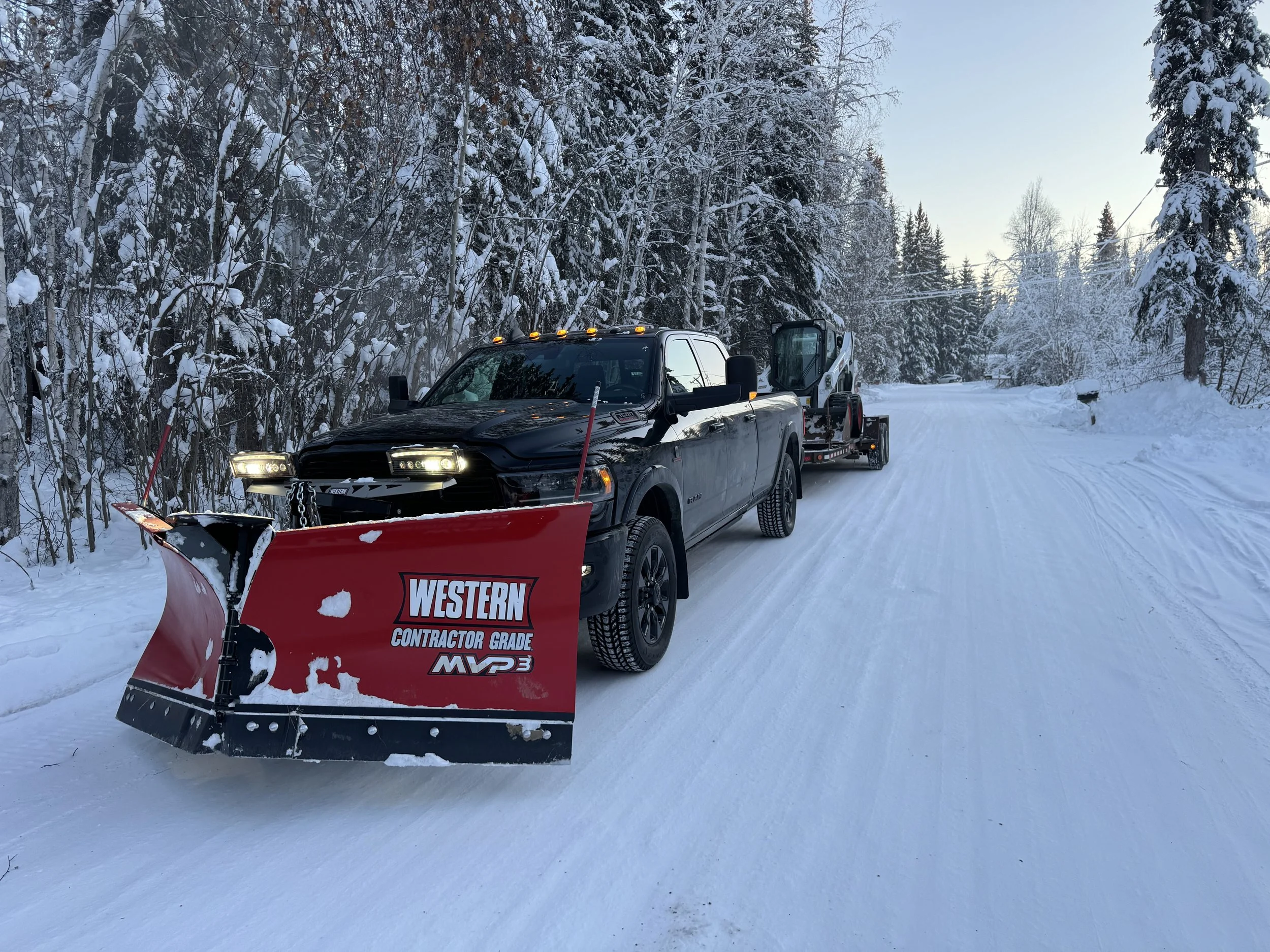 A black pickup truck equipped with a snowplow and a snow removal attachment on a snow-covered road in a winter forest setting.