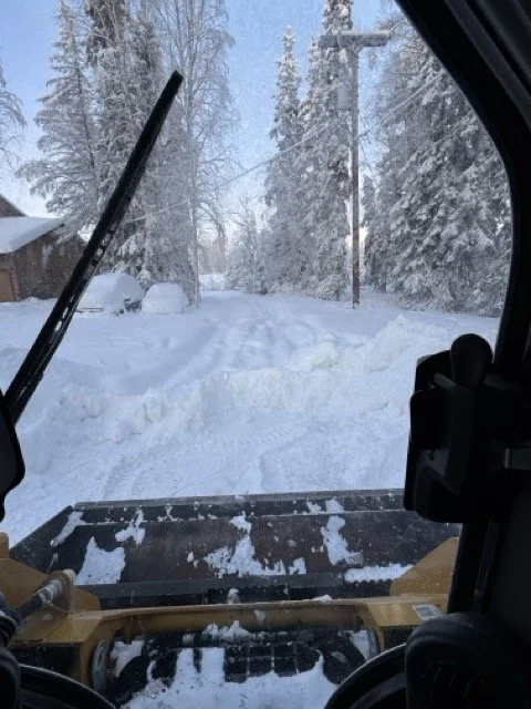 View from inside a snow removal vehicle showing a snow-covered road and tall snow-laden trees.