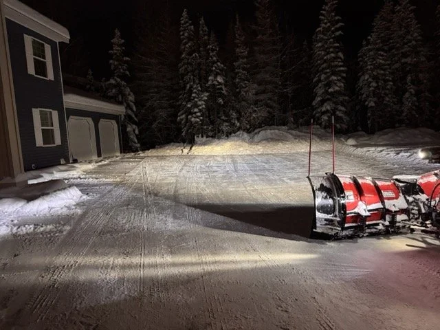 Snow-covered driveway at night with snowplow parked on the right and a house on the left, surrounded by snow-laden trees.