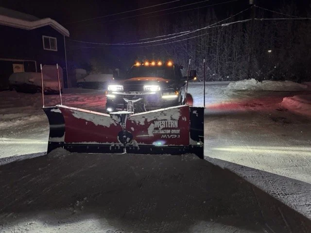 Snow-covered street at night with a red snowplow and a dark pickup truck facing the camera