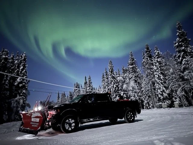 A black pickup truck with a snow plow attached to the front, parked on snow-covered ground, with snow-covered trees and the northern lights in the sky above.