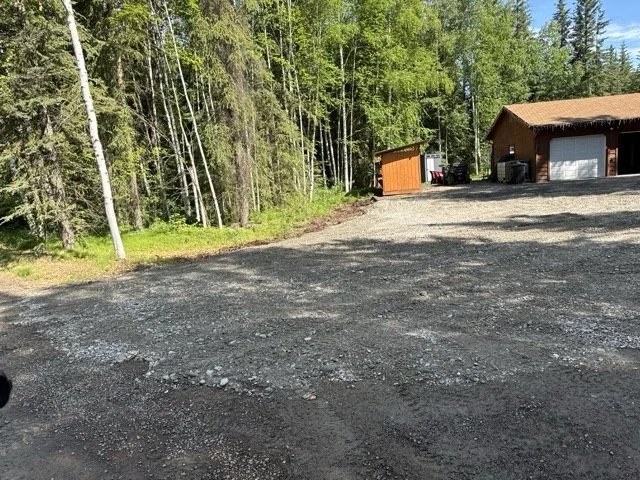 Gravel driveway leading to a wooden garage and shed, surrounded by trees and utility poles.