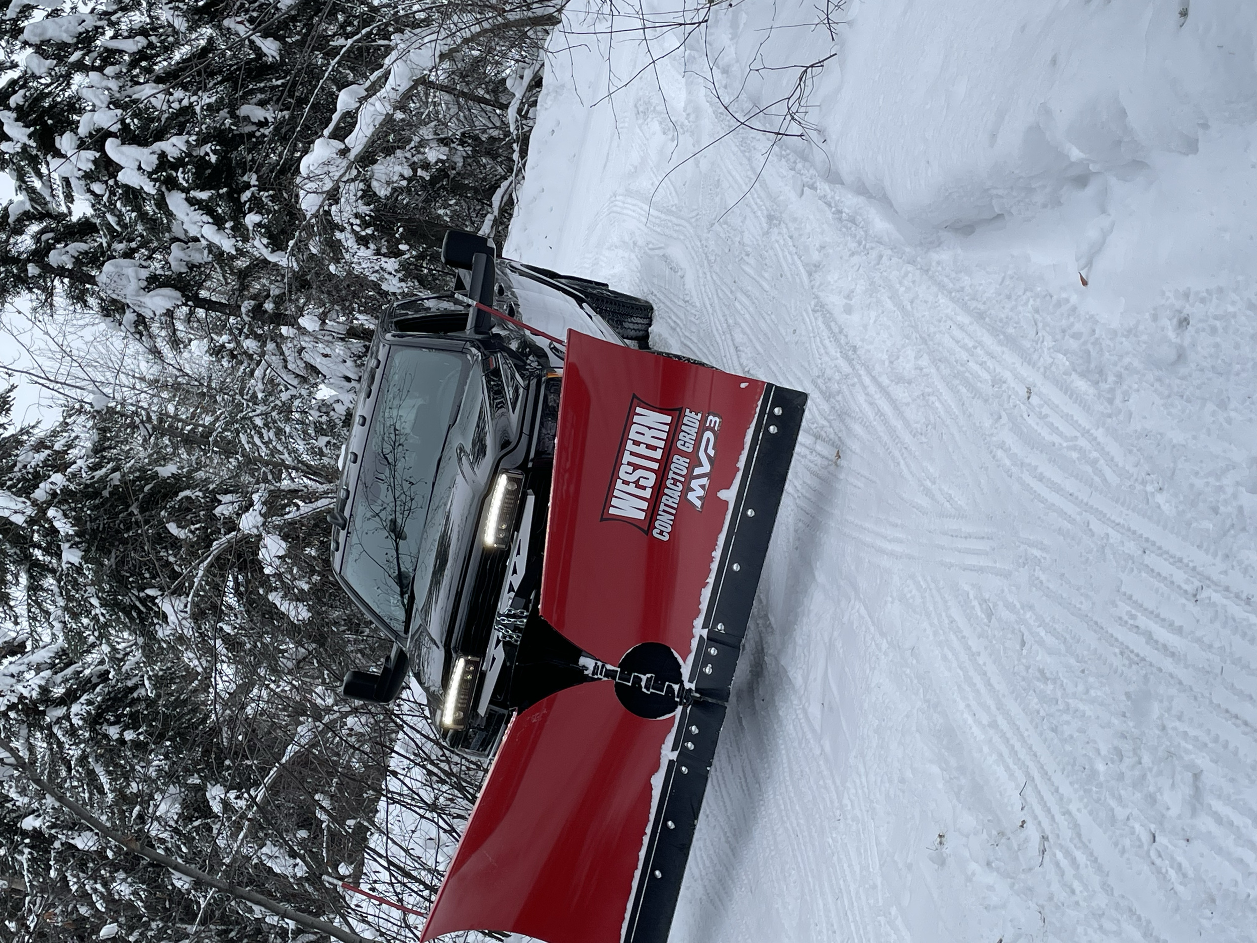 A snowplow with a red blade and black body, displaying a logo and text for Western Contracting, clearing snow on a winter trail surrounded by snow-covered trees.
