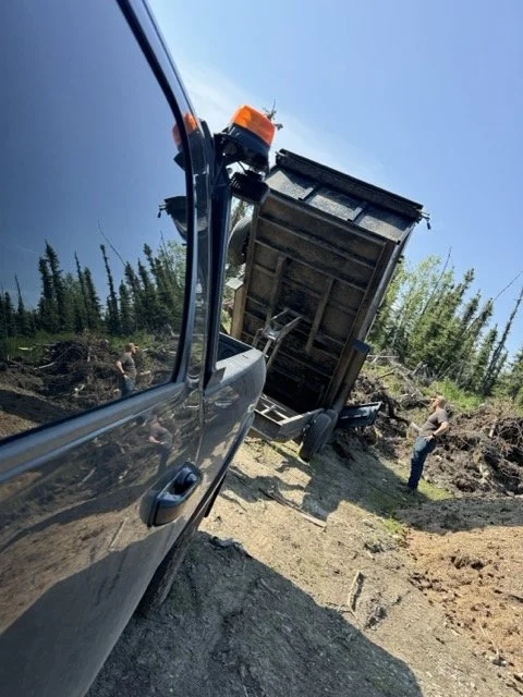 A large black dump truck tipped over on its side in a dirt area, with workers nearby and trees in the background, during daytime.