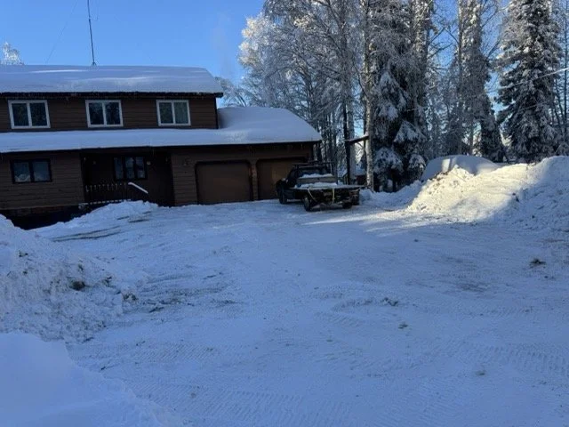 A snow-covered driveway in front of a brown house with a closed garage door, surrounded by snow and tall trees in the background.